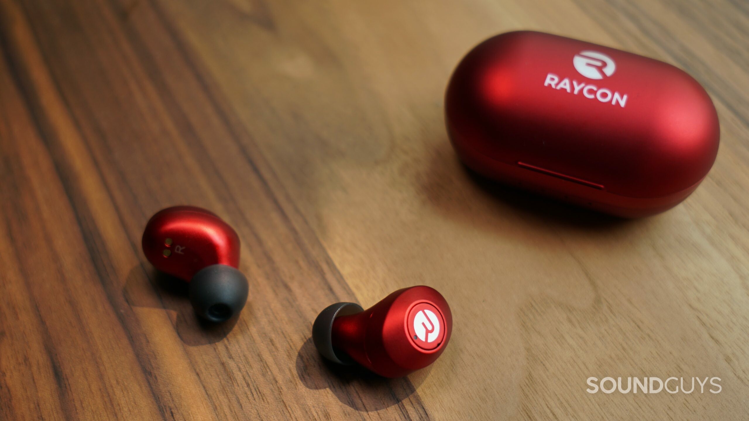 The Raycon Everyday Earbuds sits on a wooden table, with the earbuds laying next to the charging case.