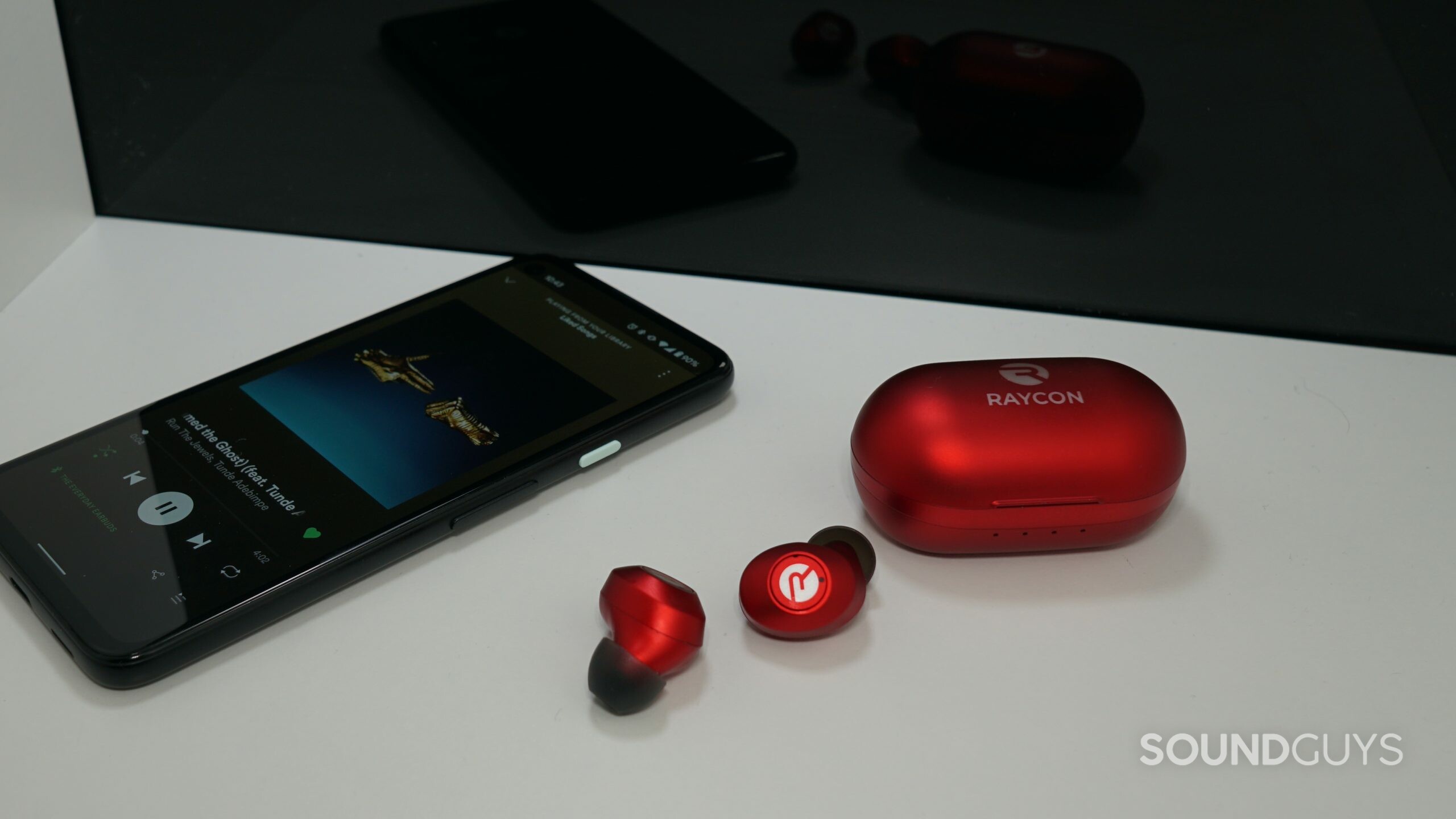 Raycon-Everyday-Earbuds-phone-shot The Raycon Everyday Earbuds lay on a white shelf in front of a reflective black surface, next to Google Pixel 4a.