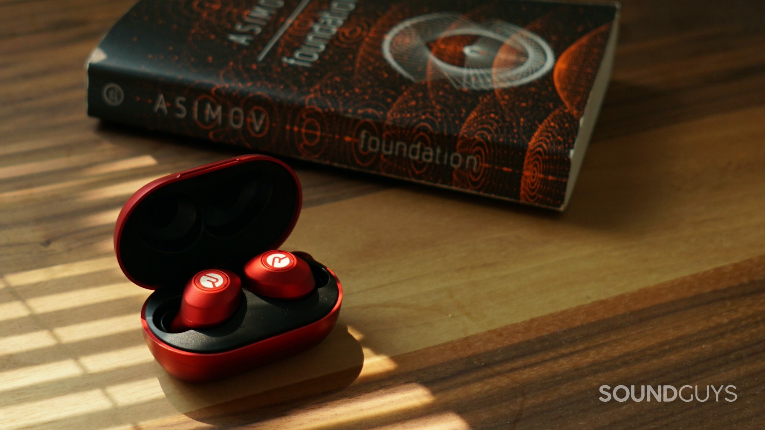 The Raycon Everyday Earbuds sit in their open charging case on a wooden table next to a copy of a Foundation by Isaac Asimov.