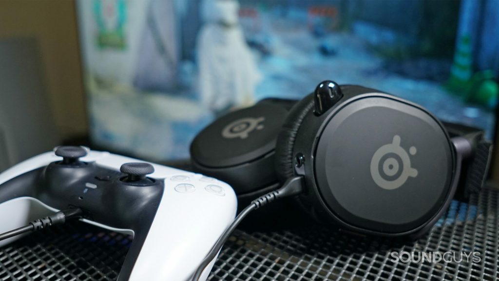 The SteelSeries Arctis Prime gaming headset lays on a metal table next to a PlayStation DualSense controller, which it's plugged into, in front a TV with Star Wars Jedi: Fallen Order running on PlayStation 5.