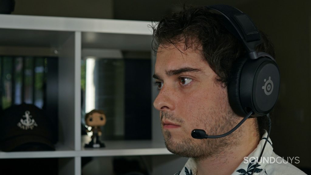 A man wears the SteelSeries Arctis Prime gaming headset seated at a desk in front of a shelf with a Han Solo Funko! Pop and a hat for the Sappyfest music festival on it.