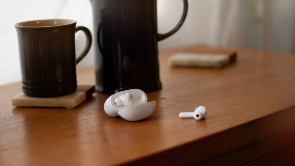 TCL MOVEAUDIO S600 case open with one earbud out on a wood table with a coffee mug.