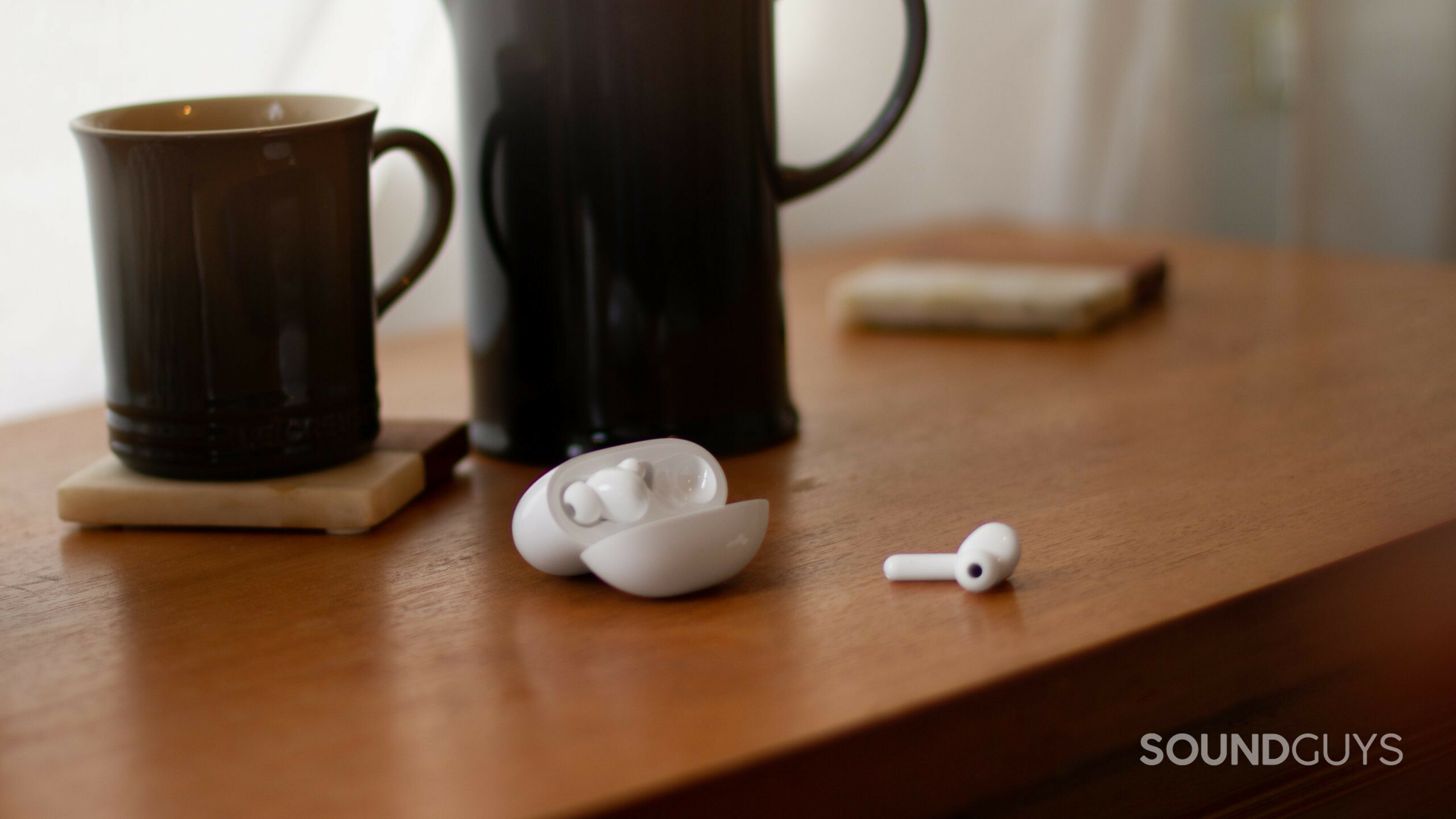 TCL MOVEAUDIO S600 case open with one earbud out on a wood table with a coffee mug.