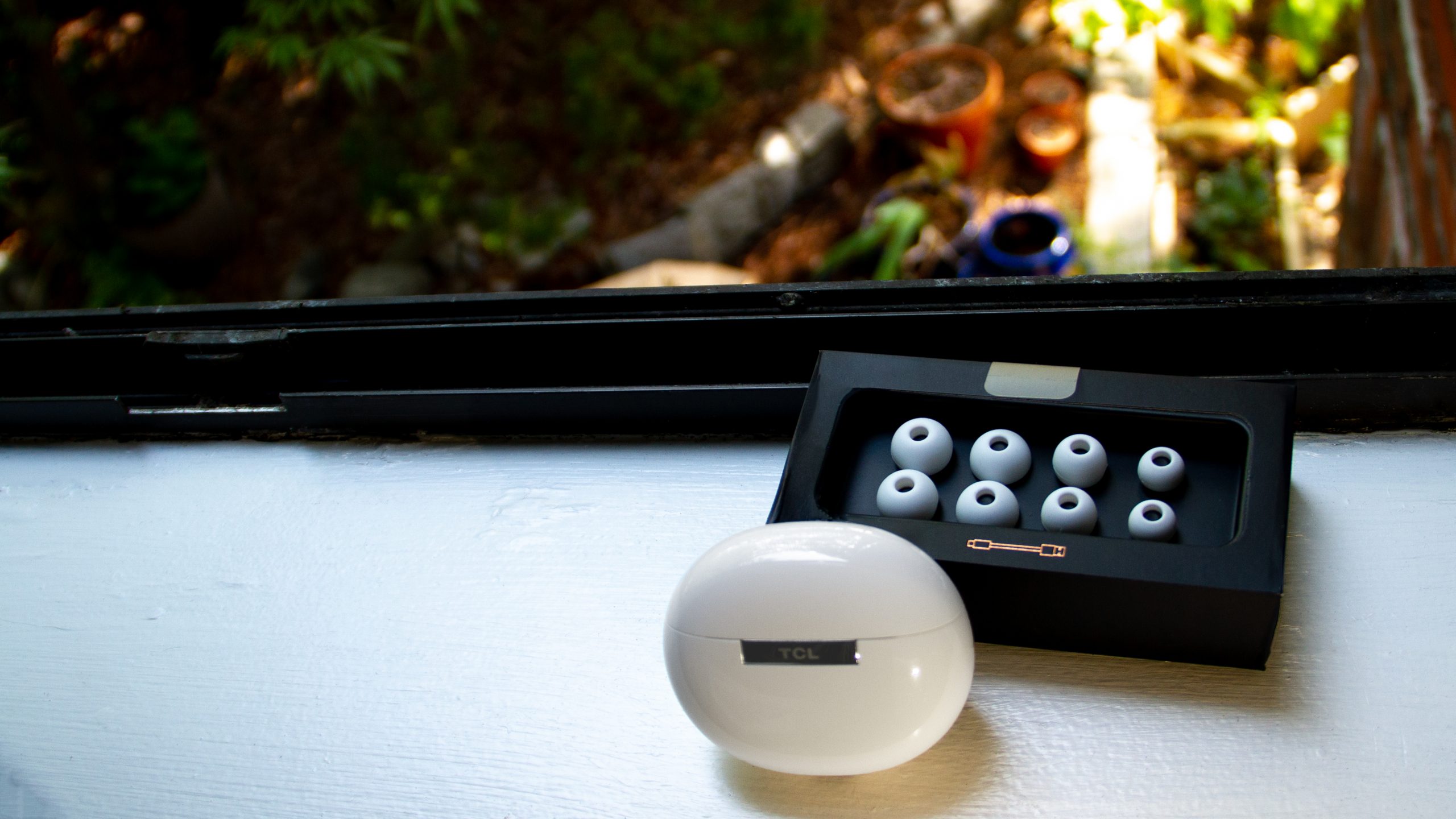 The TCL MOVEAUDIO S600 case and four additional silicone ear tips sit on a white window sill with a blurred garden in the background.