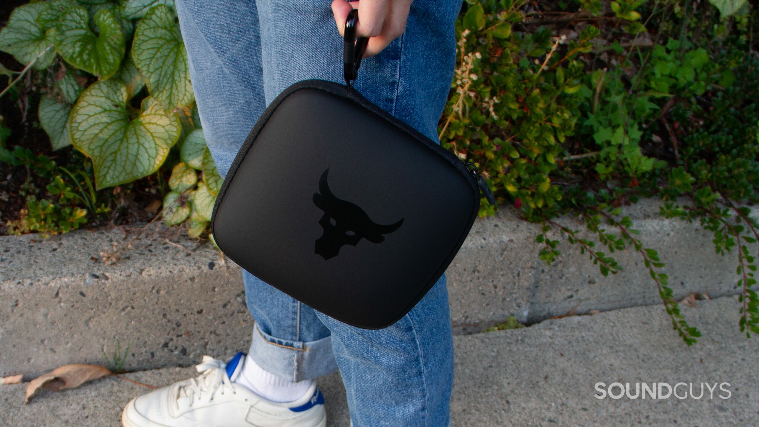 Image is of a man holding the case of the Under Armour Project Rock by JBL headphones by the carabiner by his legs while standing on cement by a green bush.