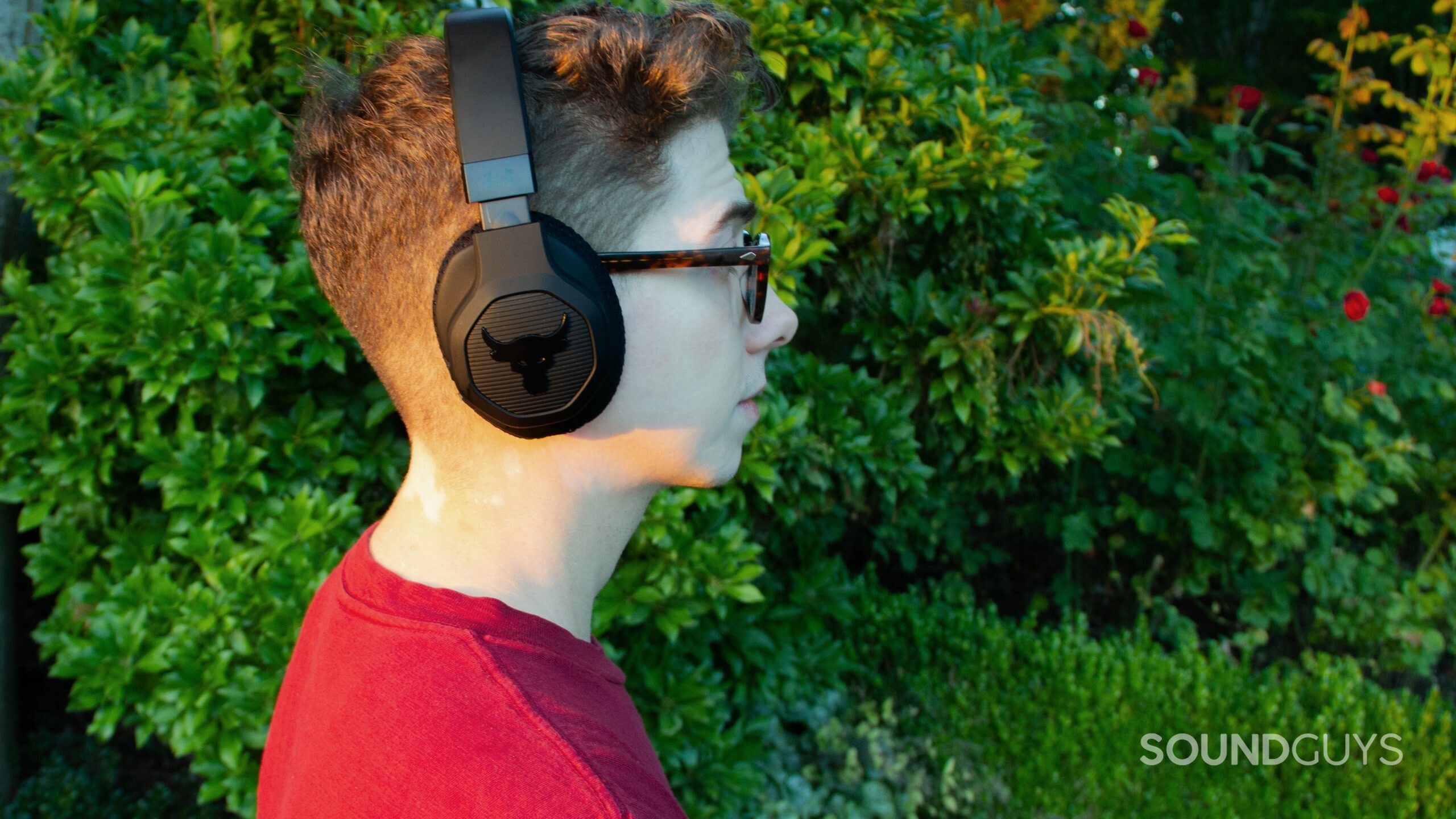 A man standing at profile wear Under Armour Project Rock by JBL headphones outside by greenery in a raspberry colored shirt.