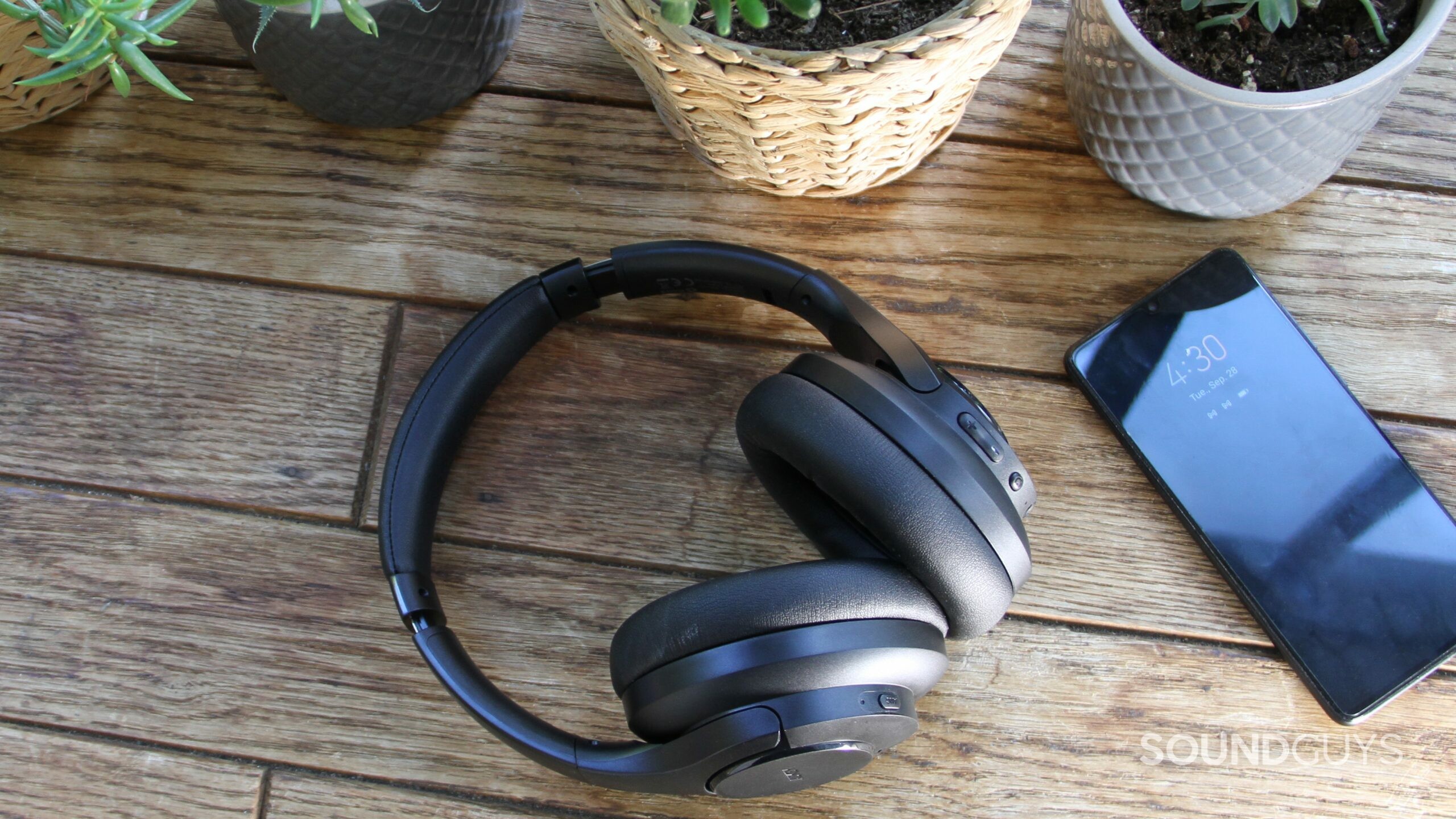 Aukey Hybrid Active Noise Canceling Headphones seen from above on a wood surface with a smartphone and potted plants