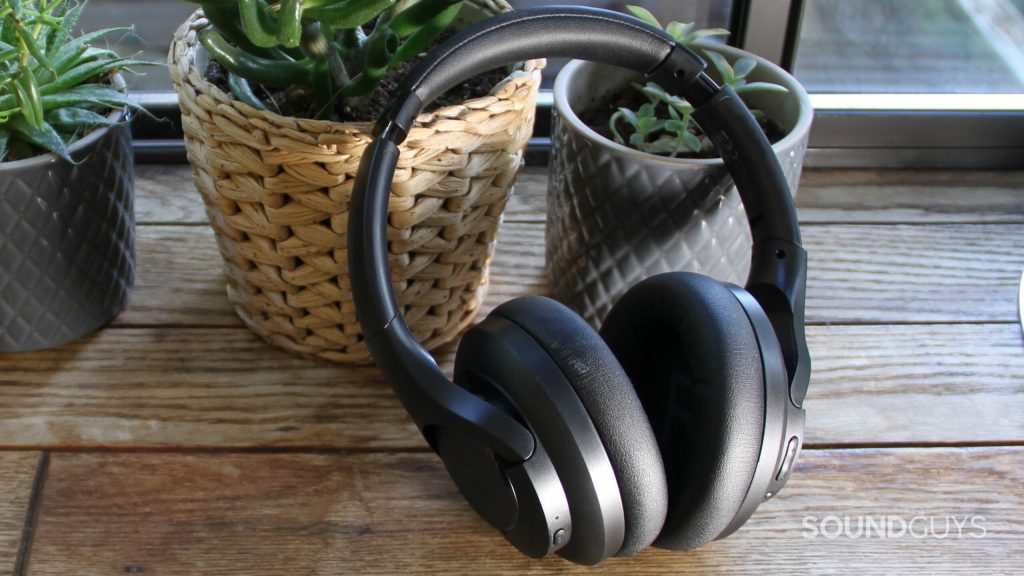 Aukey Hybrid Active Noise Cancelling Headphones leans against a grey potted plant and a wicker plant pot all sitting on a wood surface and a window in the background.