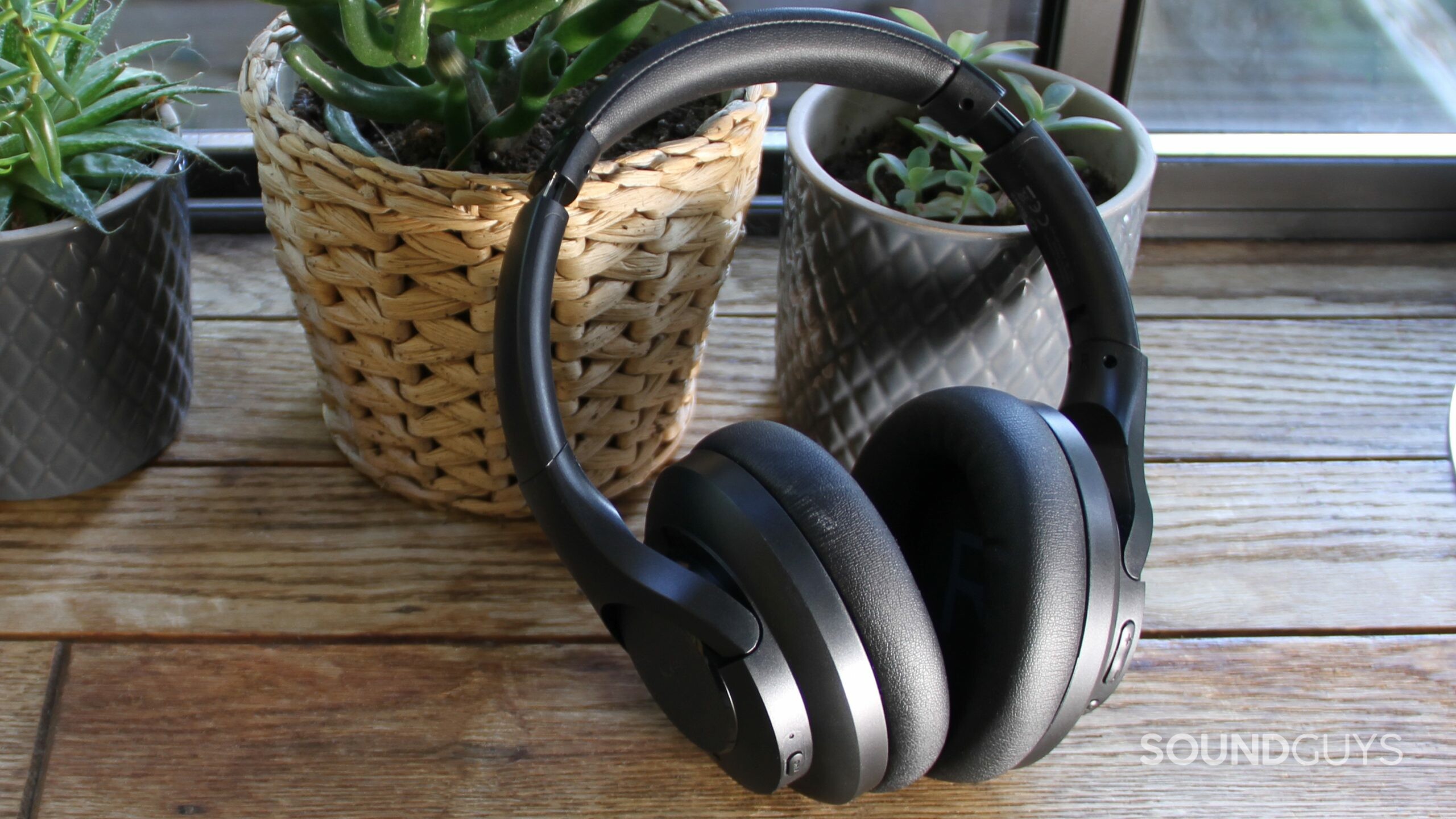 Aukey Hybrid Active Noise Canceling Headphones leans against a grey potted plant and a wicker plant pot all sitting on a wood surface and a window in the background.