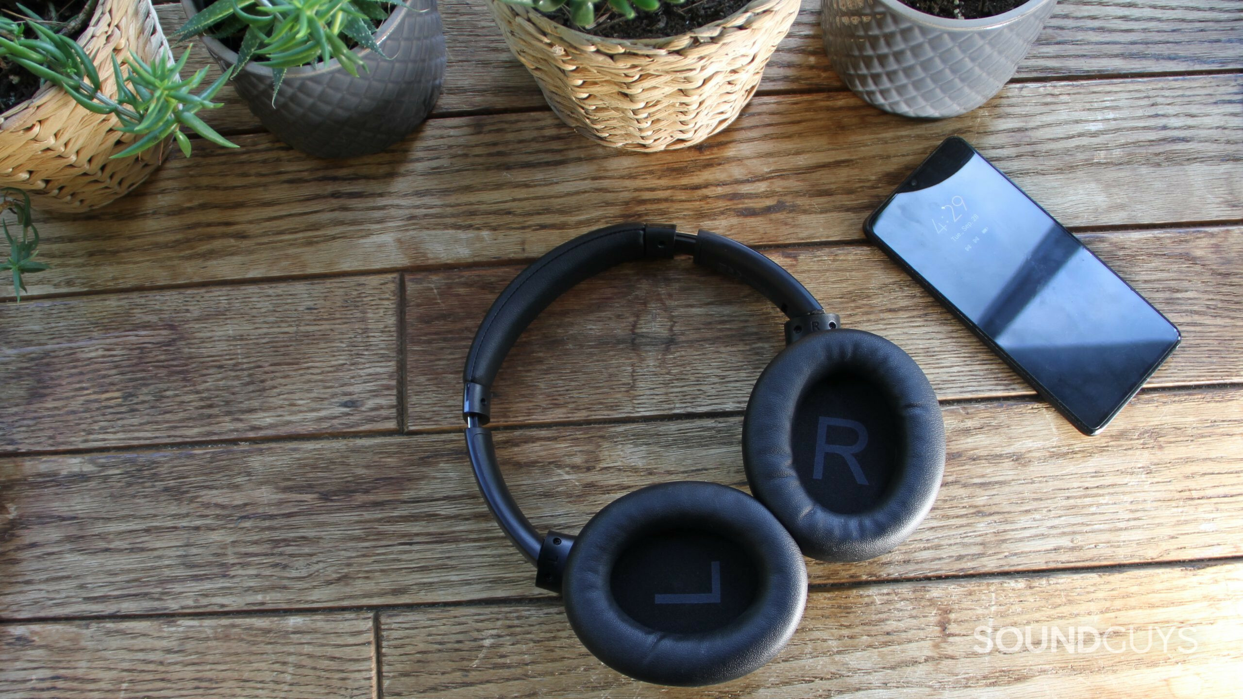 Shot from above the ear cups of the Aukey Hybrid Active Noise Canceling Headphones are folded out so you see L and R on the inside, with a smartphone and plant pots resting on a wood surface.