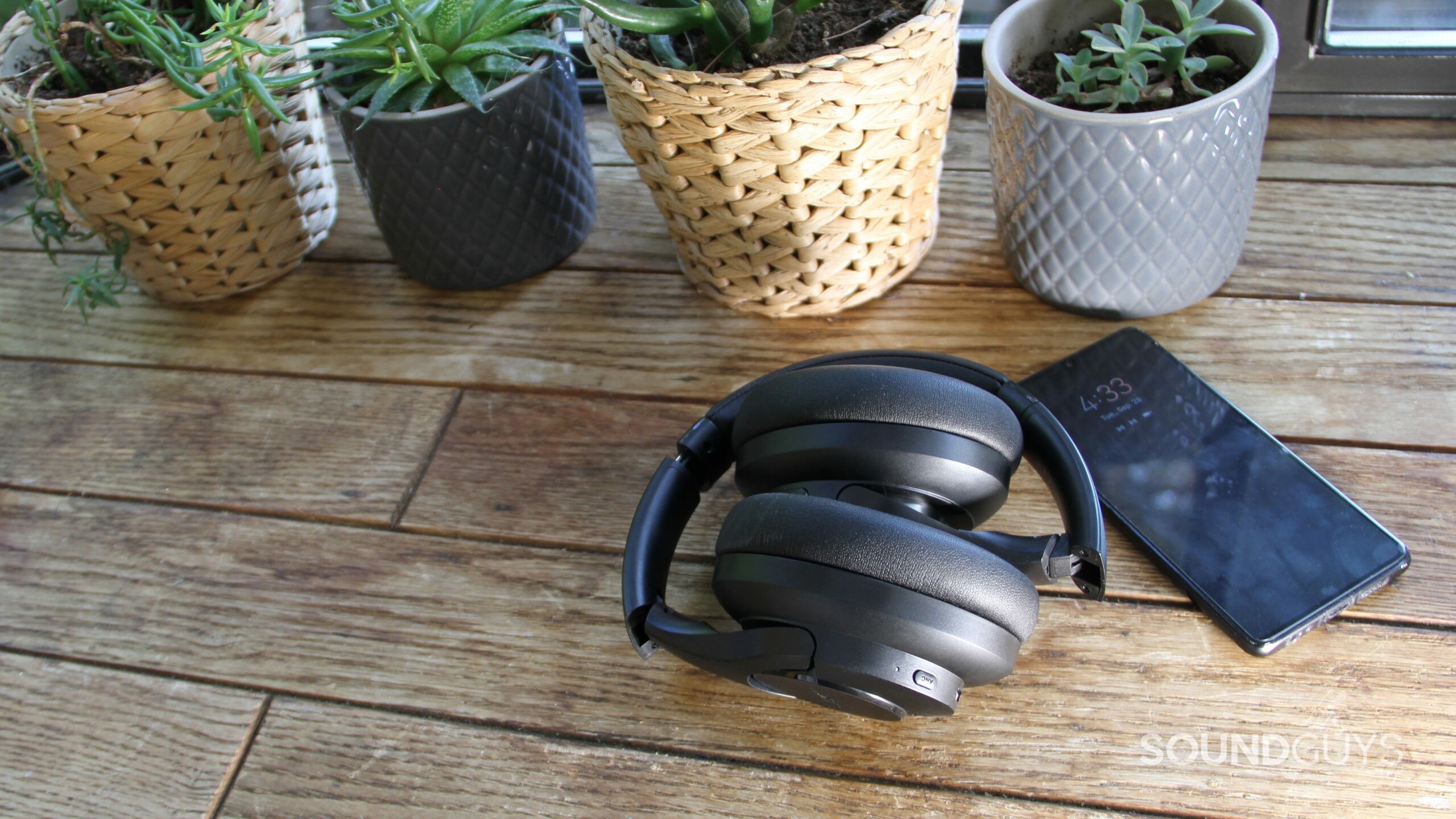 Aukey Hybrid Active Noise Canceling Headphones folded up with a smartphone and potted plants.