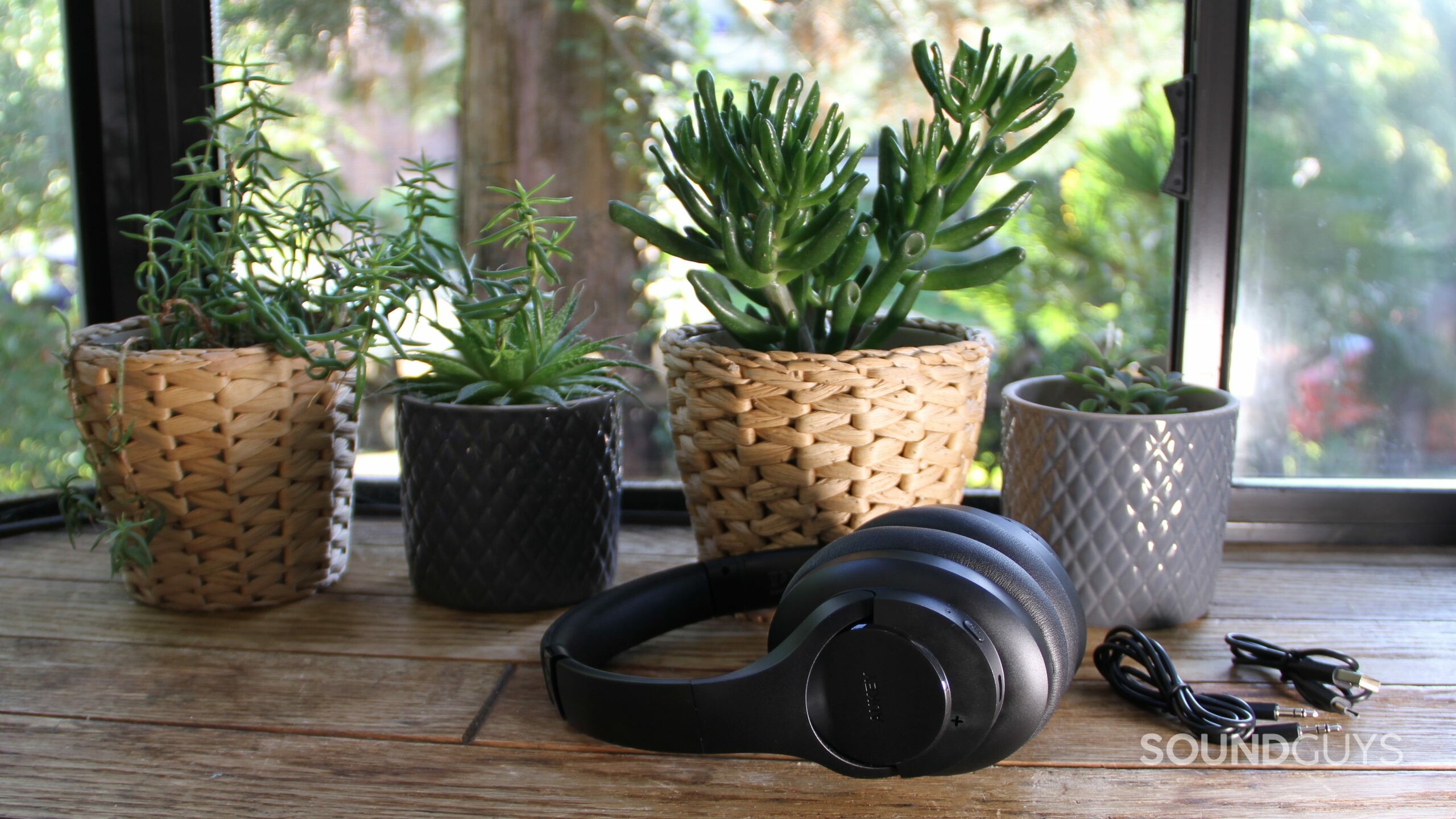 Aukey Hybrid Active Noise Canceling Headphones rests on its side with included headphone jack cable and USB-C cable in front of plants and a window.