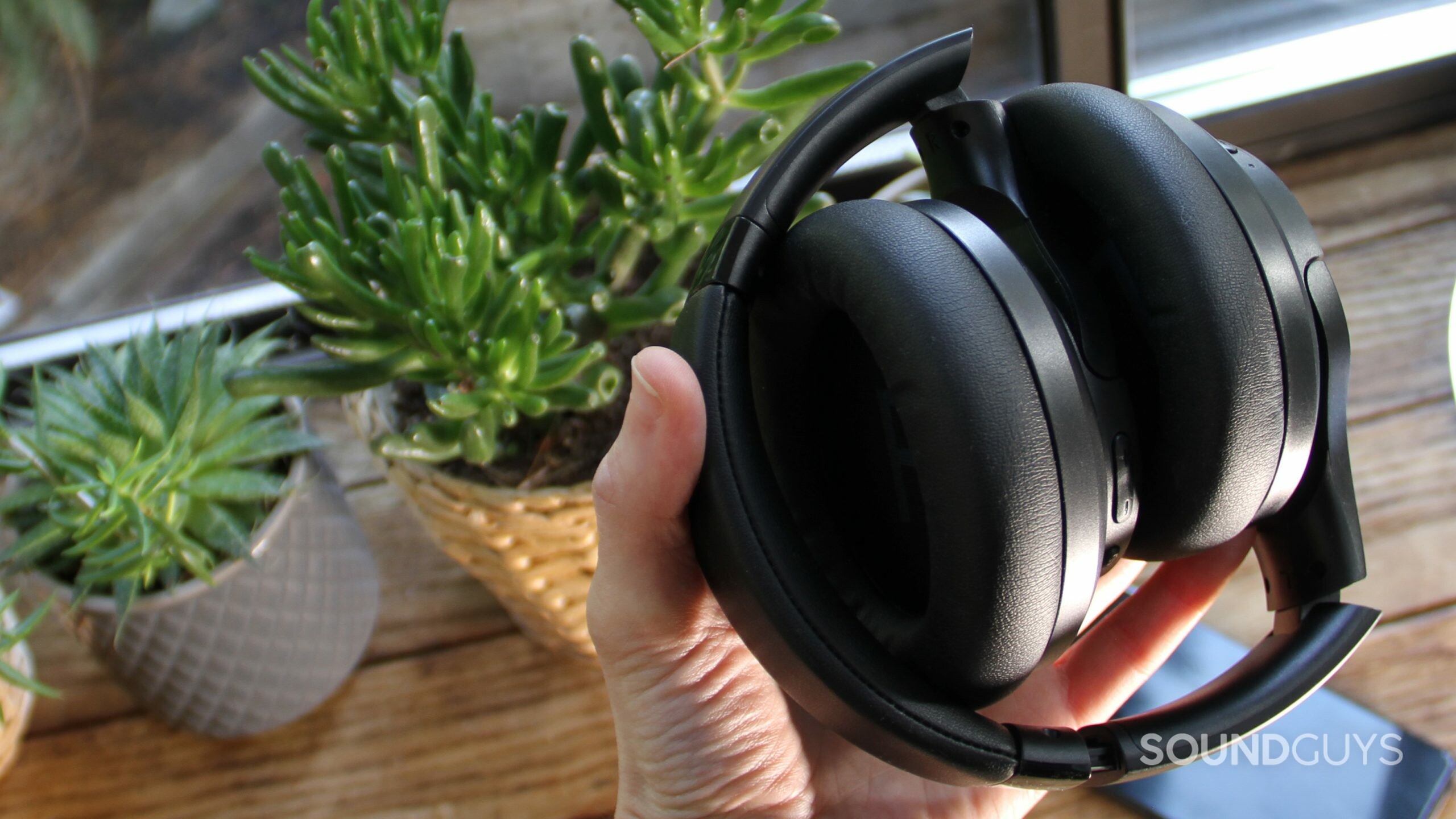 A hand holds the folded up Aukey Hybrid Active Noise Canceling Headphones with plants and a smartphone in the background.