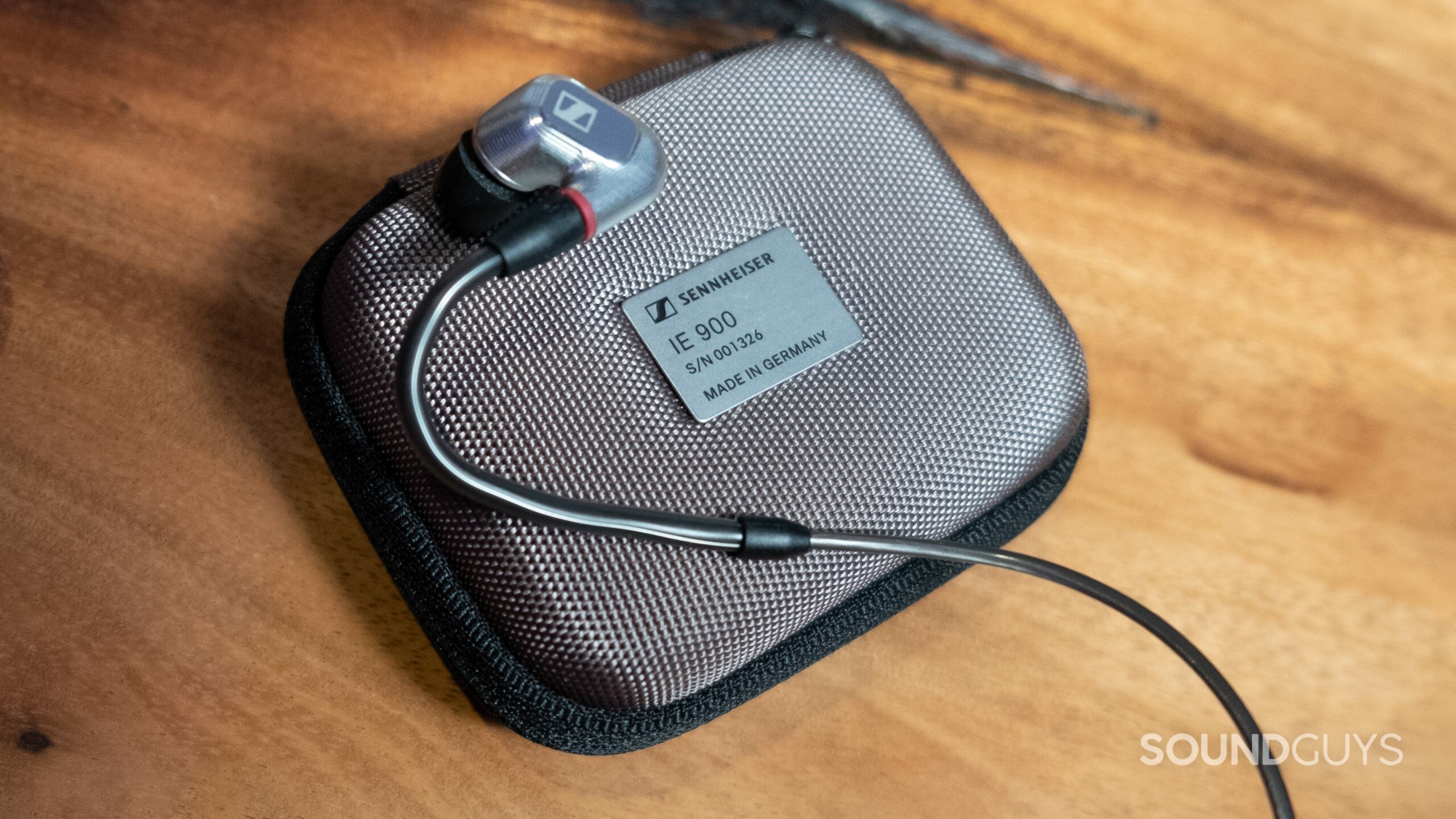 The image shows the right Sennheiser IE 900 earbud resting on top of the greyish case with individual serial number, atop a wood desk.