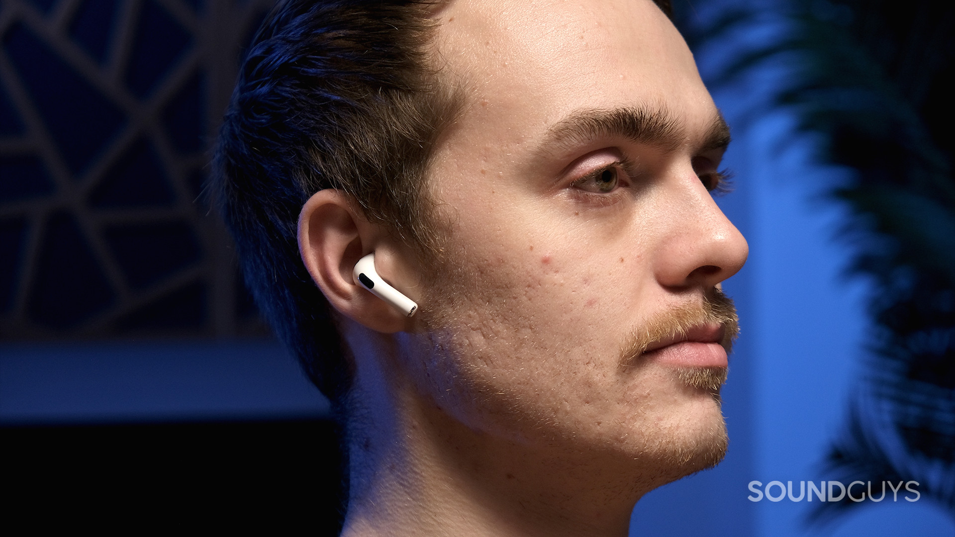 A masculine person wears the Apple AirPods (3rd generation) against a blue background.