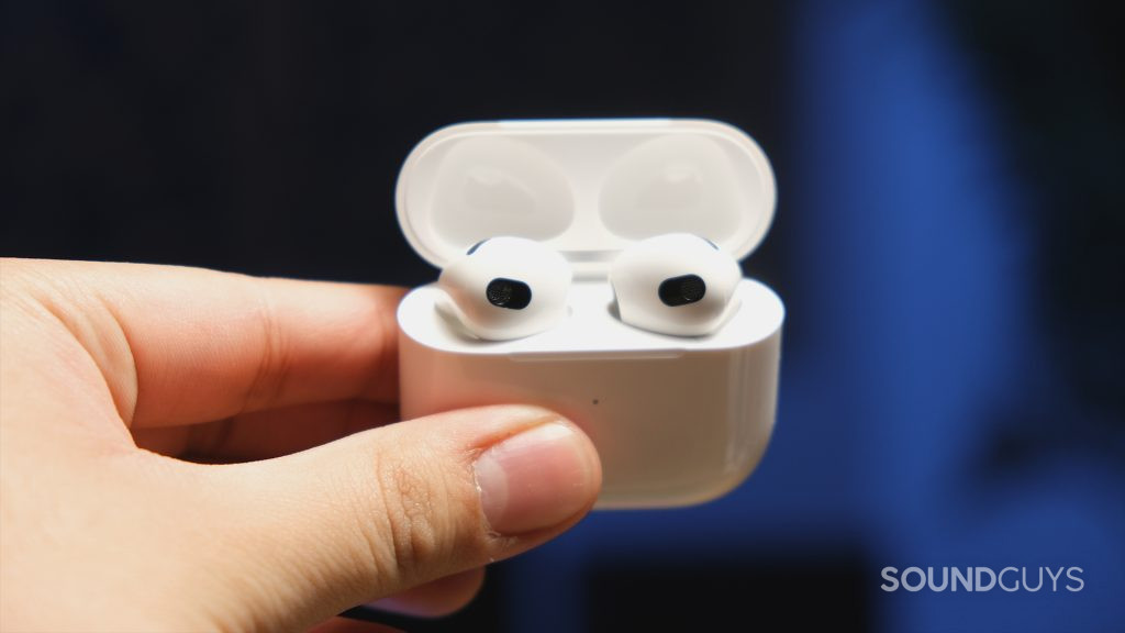 A hand holds a Apple AirPods (3rd generation) case with the earbuds in place and embedded sensors in view.