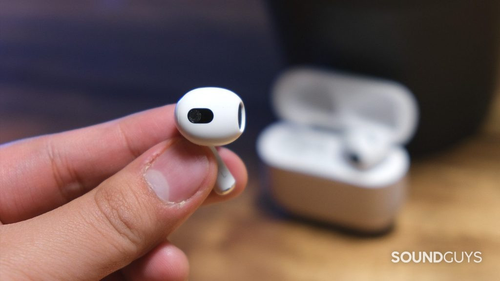 A hand holds a Apple AirPods (3rd generation) earbud by the stem to reveal the open-type fit and embedded sensors with the open case in the background.