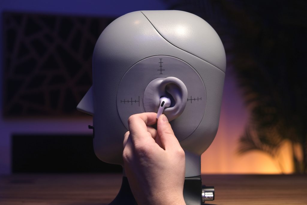 A hand holds one of the Apple AirPods (3rd generation) to the ear of a head simulator.