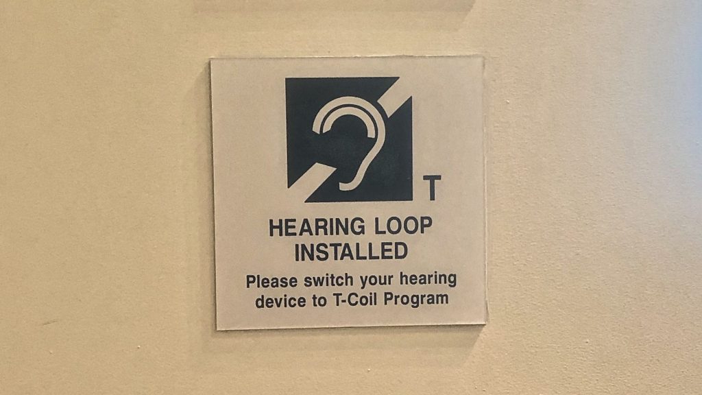 A sign outside an auditorium reads, "Hearing Loop Installed, Please switch your hearing device to T-Coil Program."
