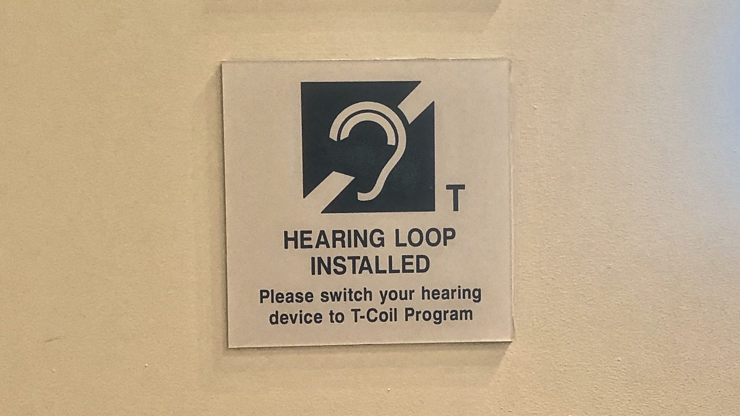 A sign outside an auditorium reads, &quot;Hearing Loop Installed, Please switch your hearing device to T-Coil Program.&quot;