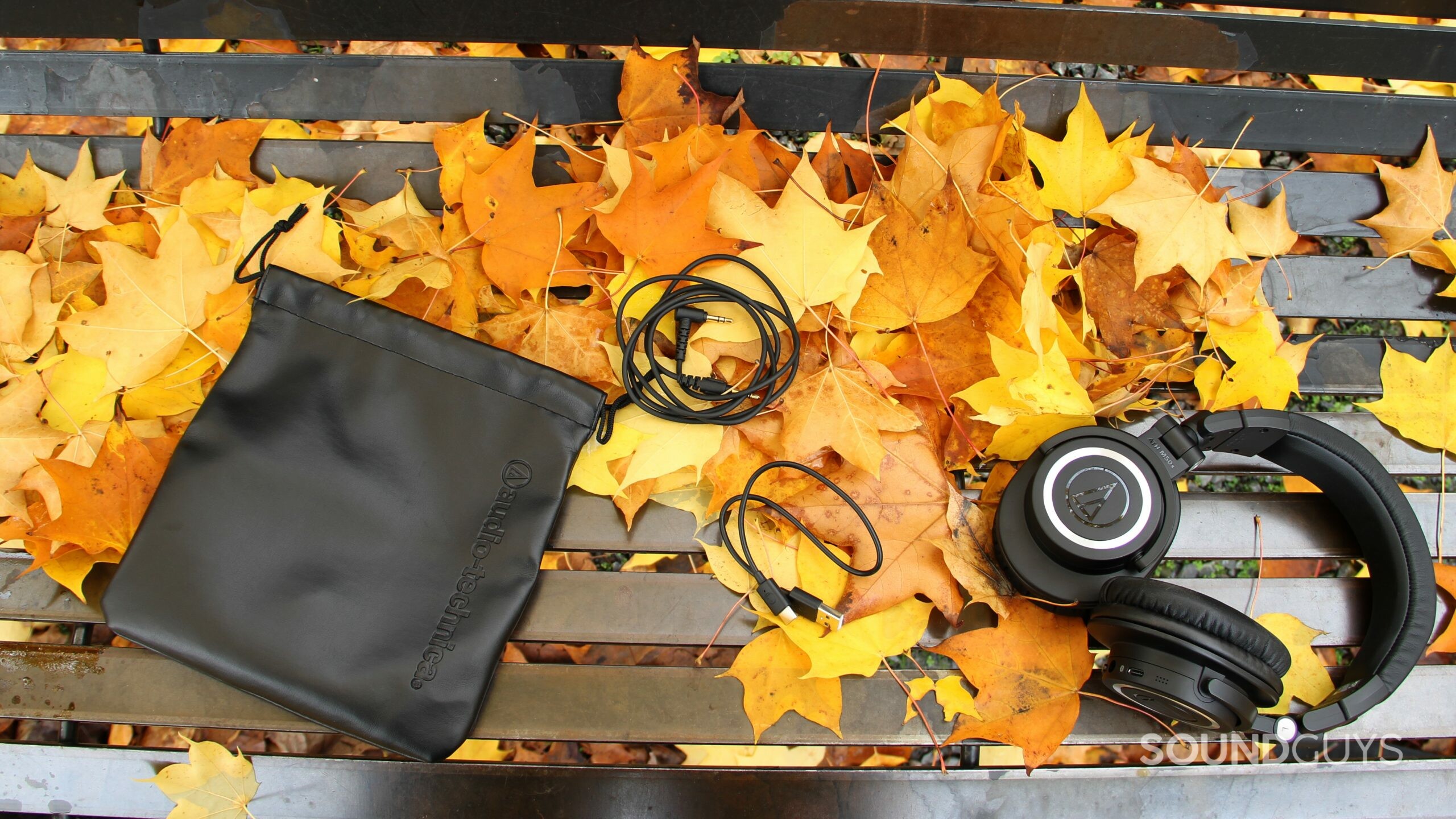 Birdseye view of Audio-Technica ATH-M50xBT2 with pouch, headphone jack, and USB cable on bench of leaves.