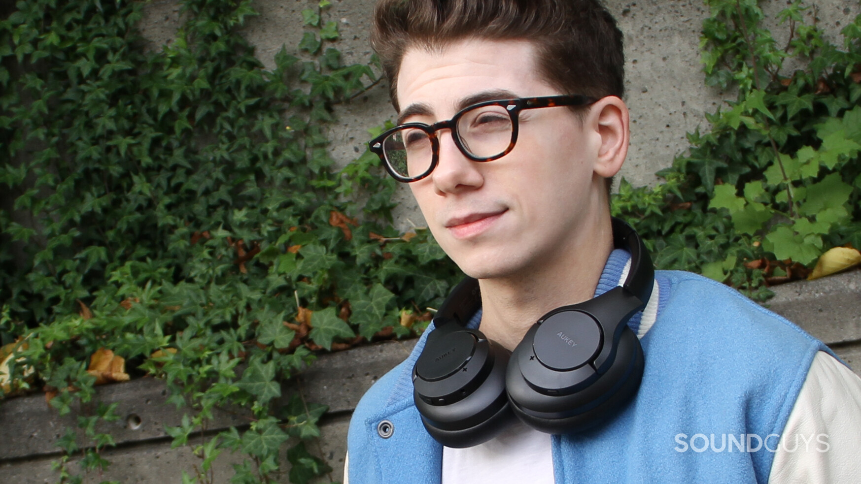 A man stands outside in front of ivy wearing the Aukey Active Noise Canceling Wireless Headphones around his neck.