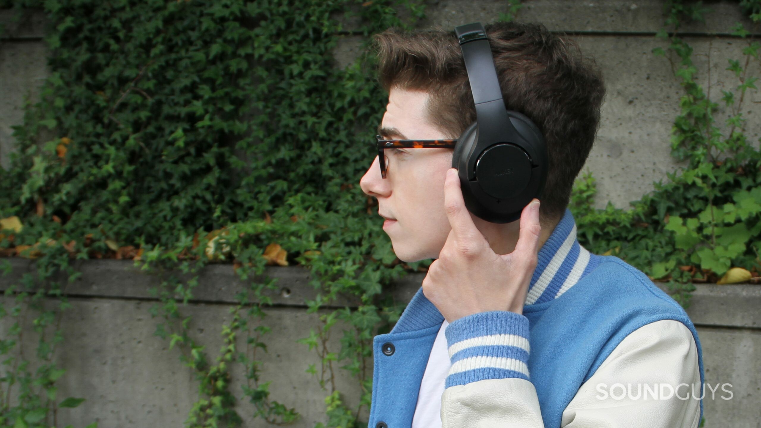 A man stands facing left outside in front of ivy and concrete wearing the Aukey Active Noise Canceling Wireless Headphones, pressing a button on the ear cup.