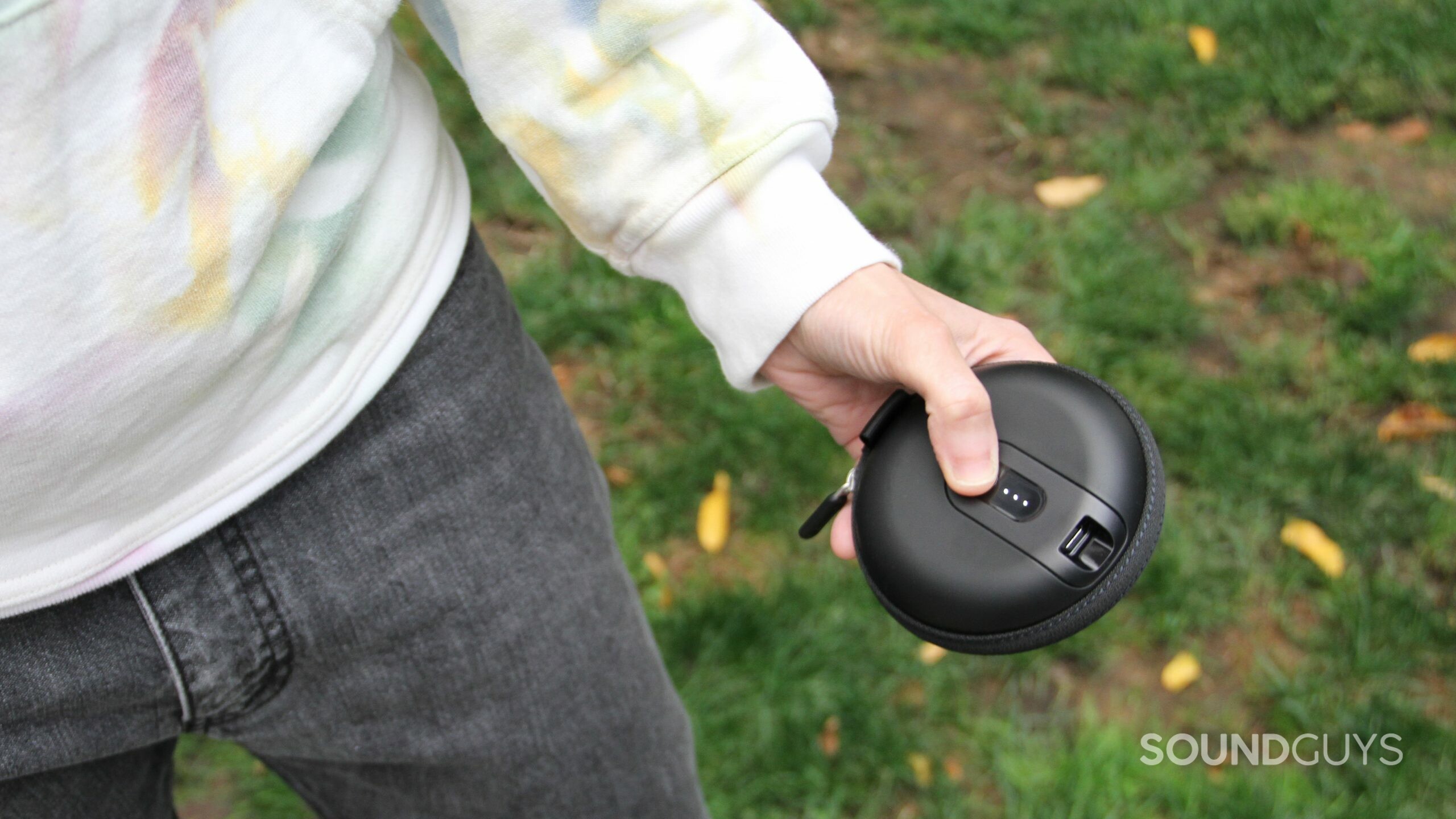 A hand holds the closed case of the Shure AONIC 215 Gen 2 with grass in the background and the battery light illuminated.