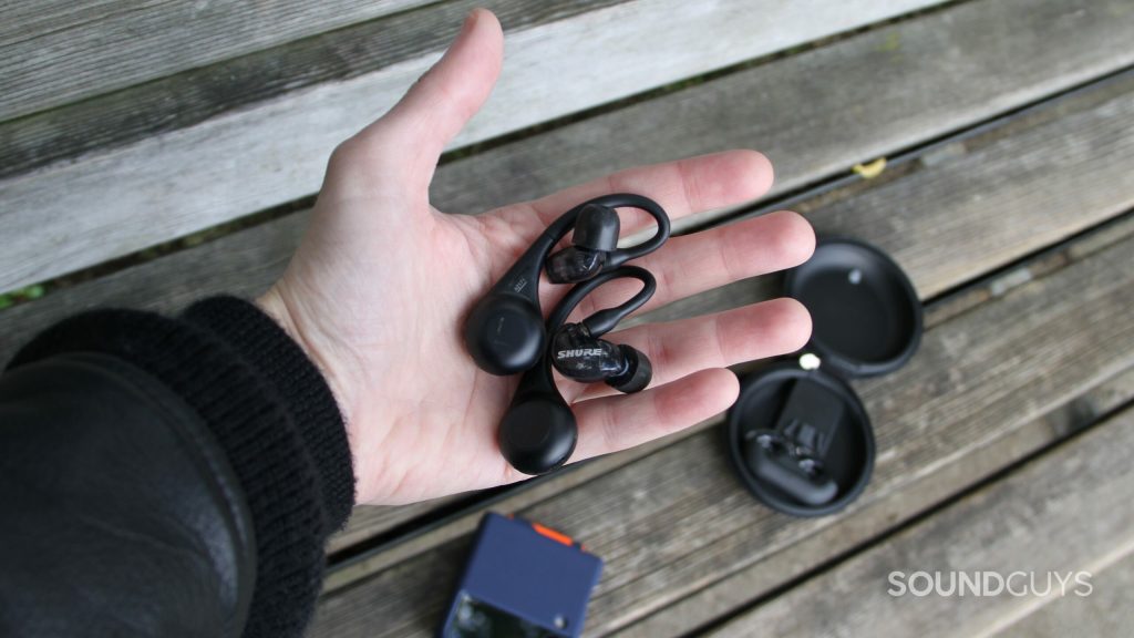 Shure AONIC 215 Gen 2 in a hand with a phone and the battery case in the background on a wood bench.