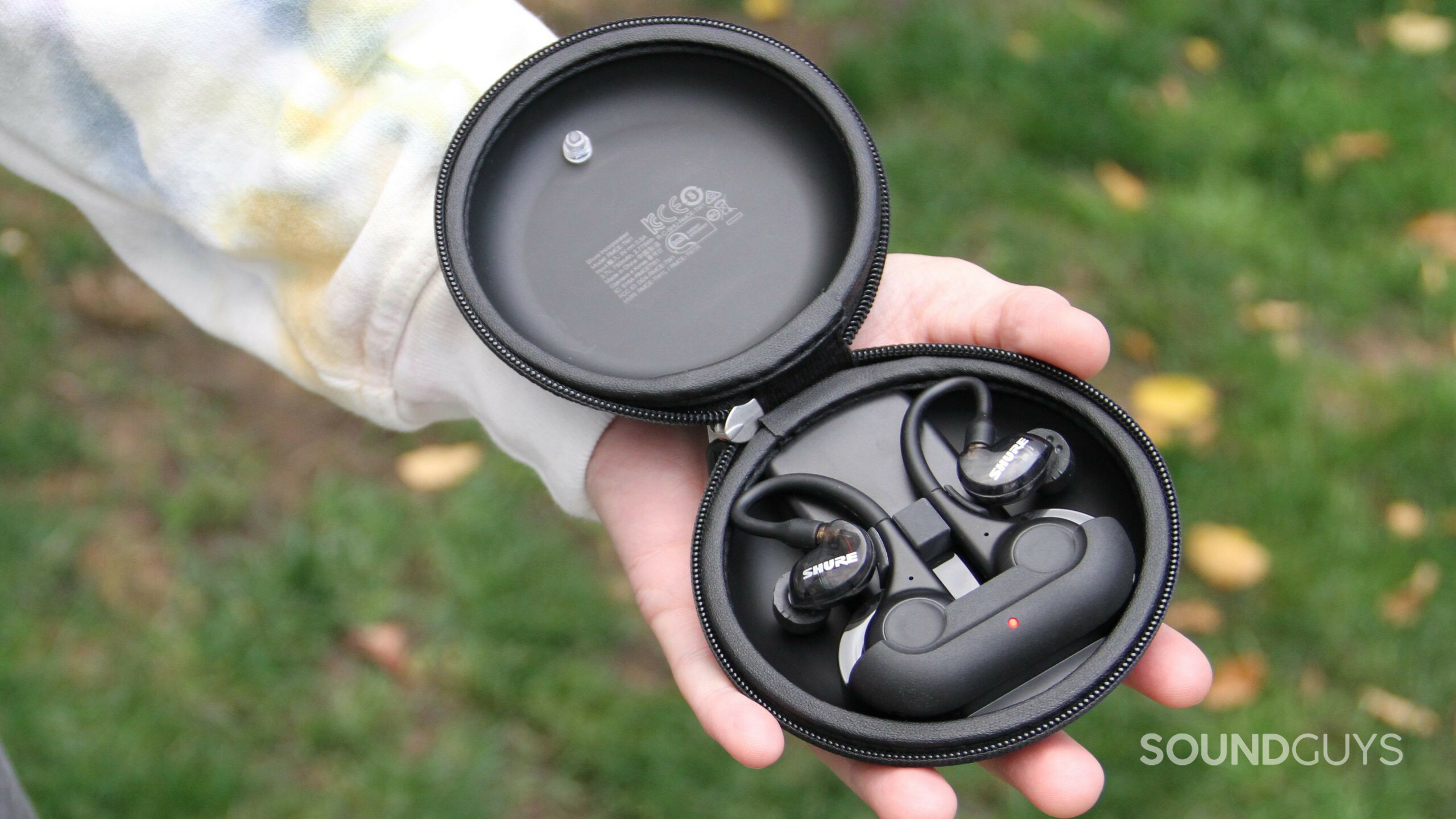 A hand holds the open battery case with the Shure AONIC 215 Gen 2 earphones inside.