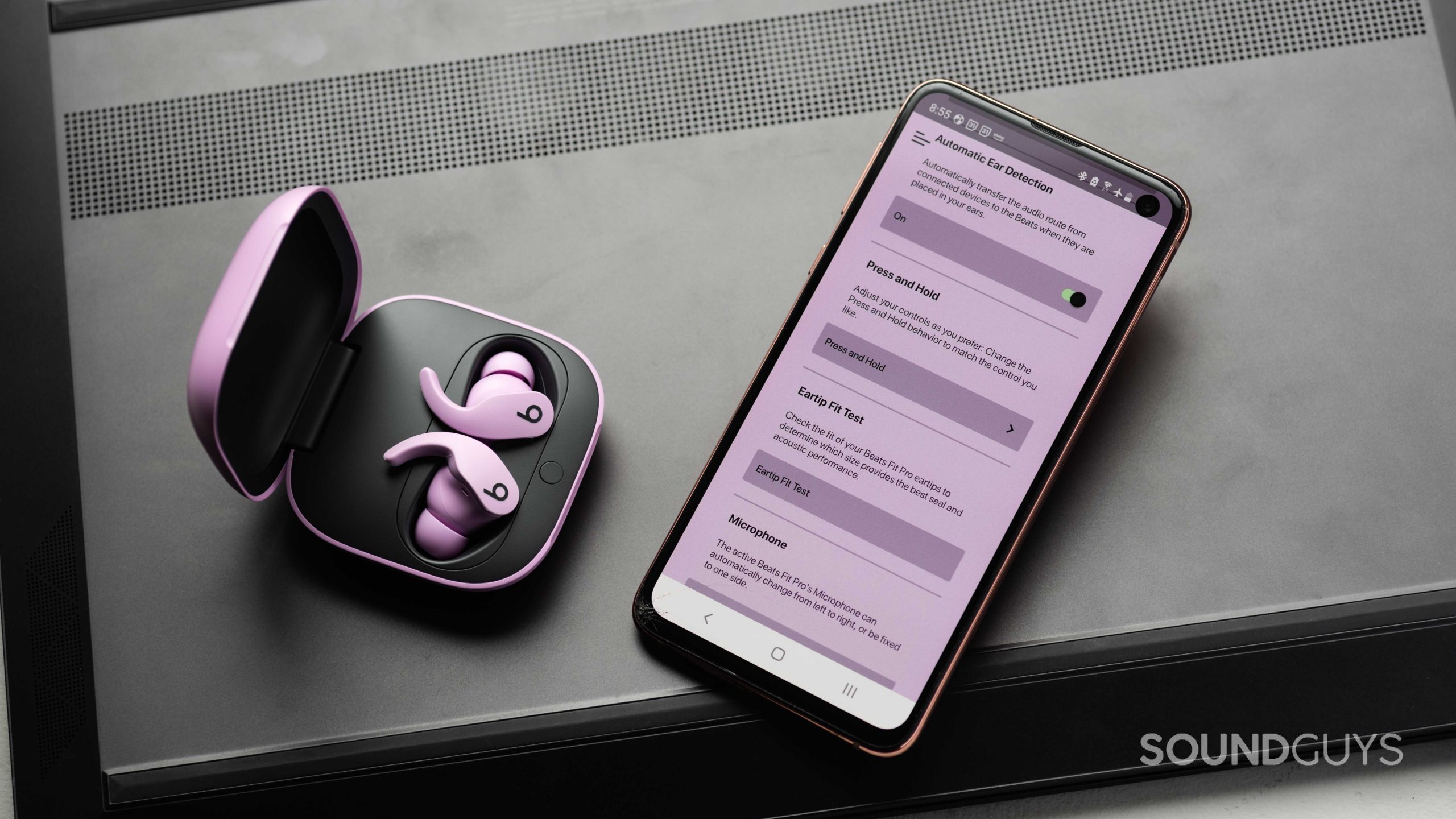 The Beats Fit Pro noise canceling true wireless earbuds in the open charging case and next to a Samsung Galaxy S10e with the Beats app open. The app has a purple tint to it, presumably to match the earphones.