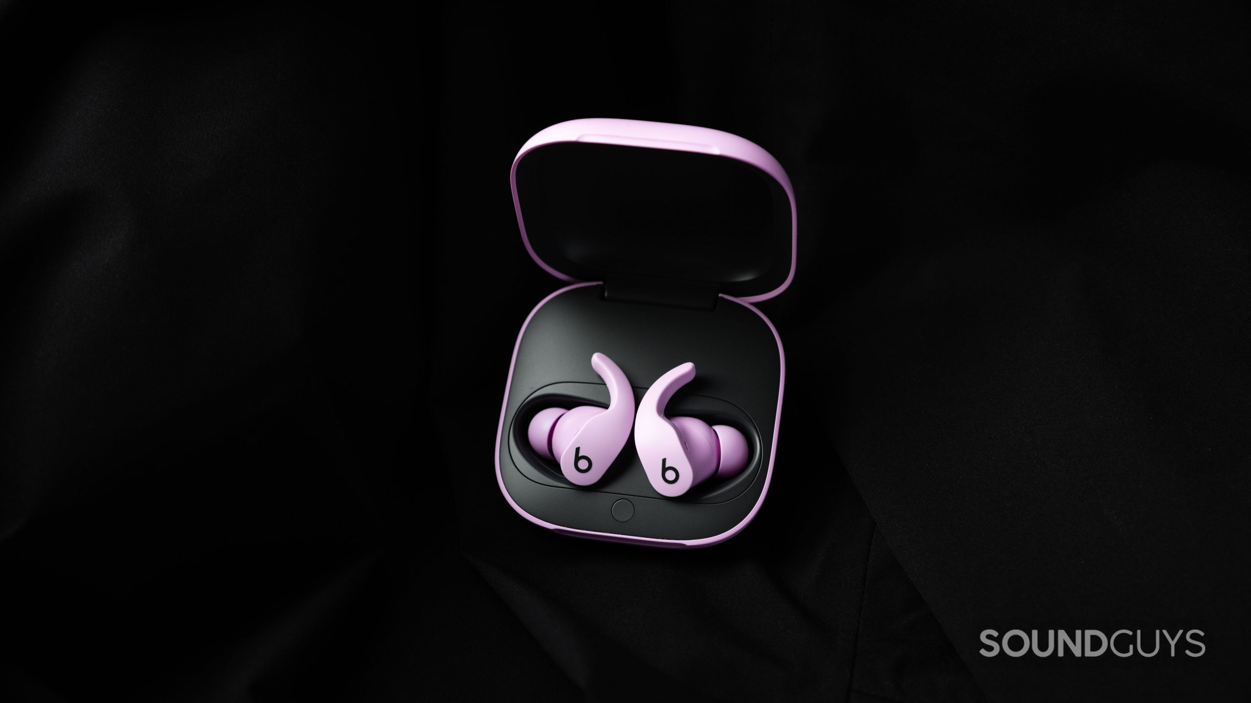 The Beats Fit Pro noise canceling true wireless earbuds in purple lay in the open charging case against a black fabric background.