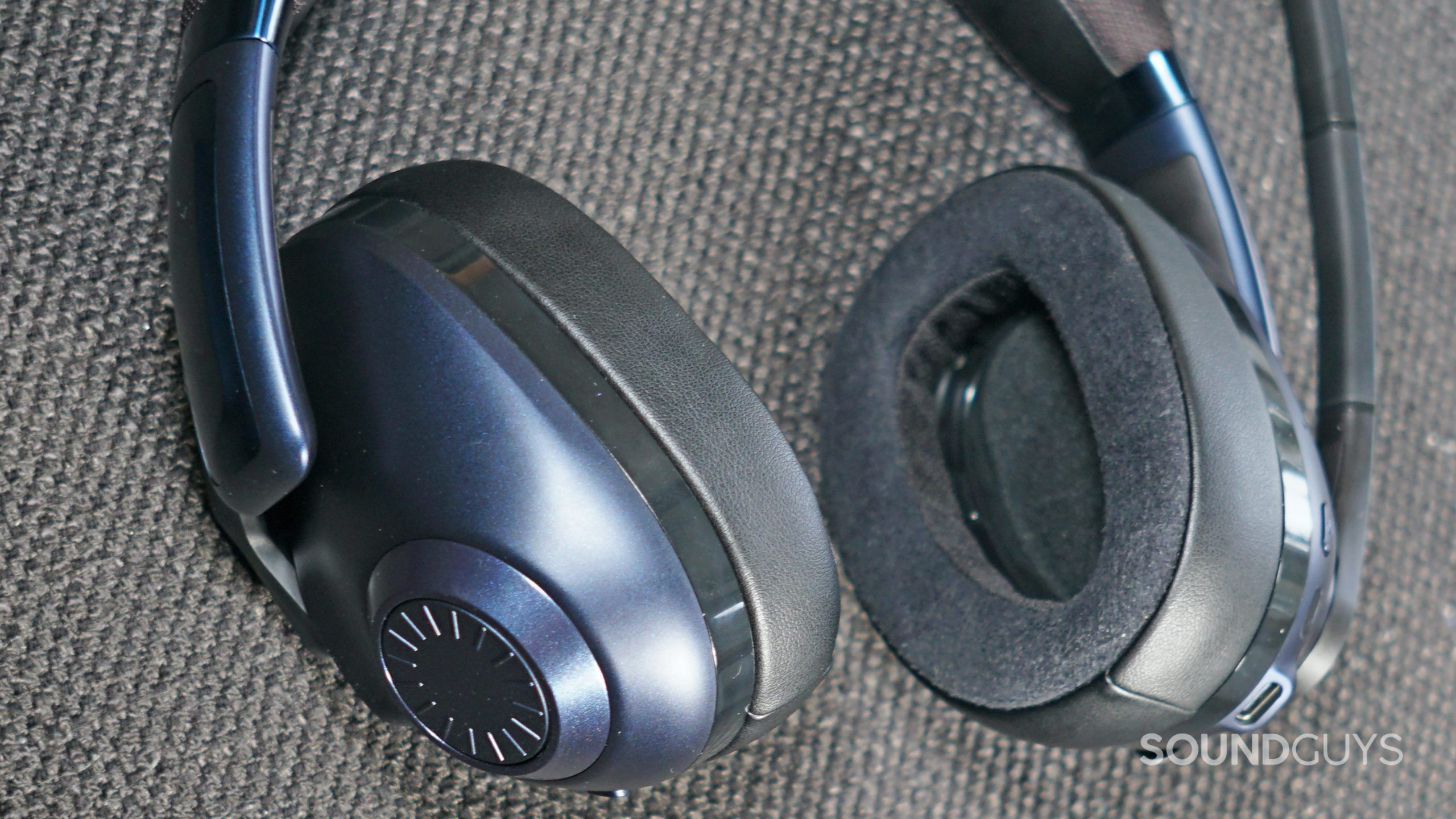 EPOS-H3PRO-Hybrid-ear-pads-and-volume The EPOS H3PRO Hybrid lays on a fabric surface with its ear pads and volume dial in view.