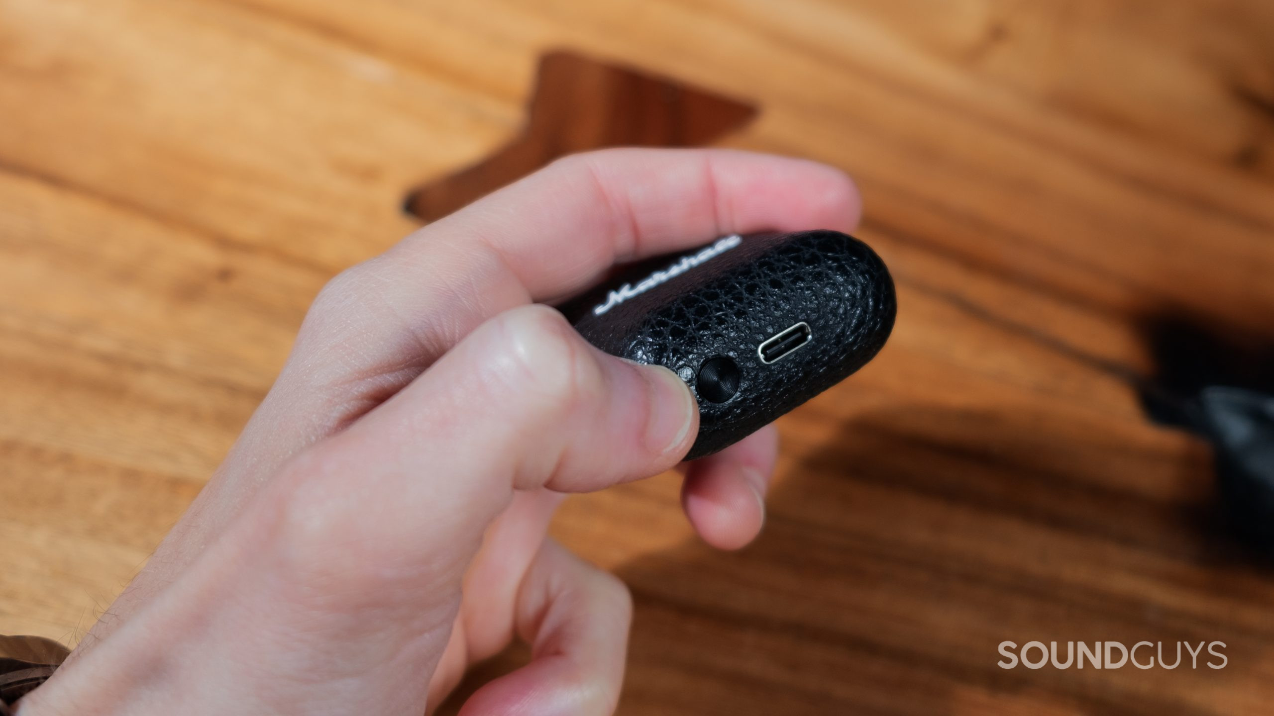 A hand holds the Marshall Minor III case showing the pairing button and USB-C connector.