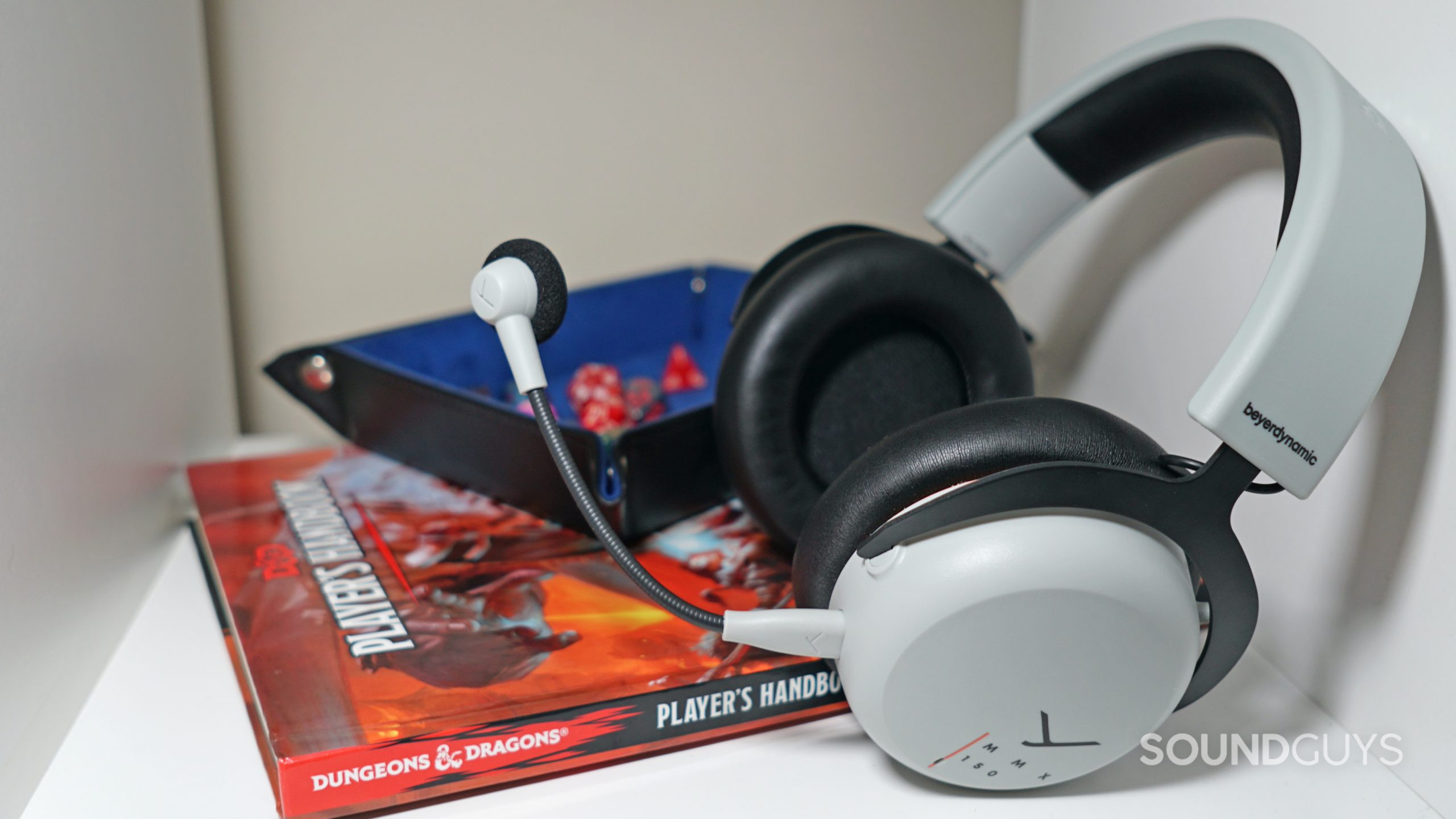 The Beyerdynamic MMX 150 lays on a white shelf next to a copy of the Dungeons and Dragons 5th edition Player's Handbook and some polyhedral dice.