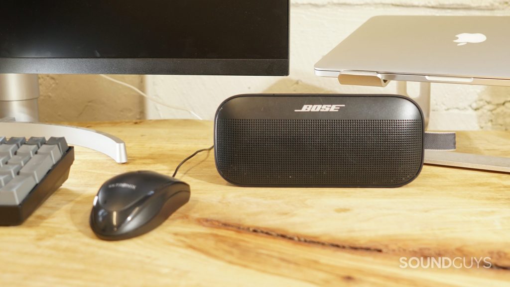 A photo of the Bose SoundLink Flex Bluetooth speaker sitting on a desk next to a monitor and an Apple MacBook Pro visible in the background and a mouse and keyboard to the left.