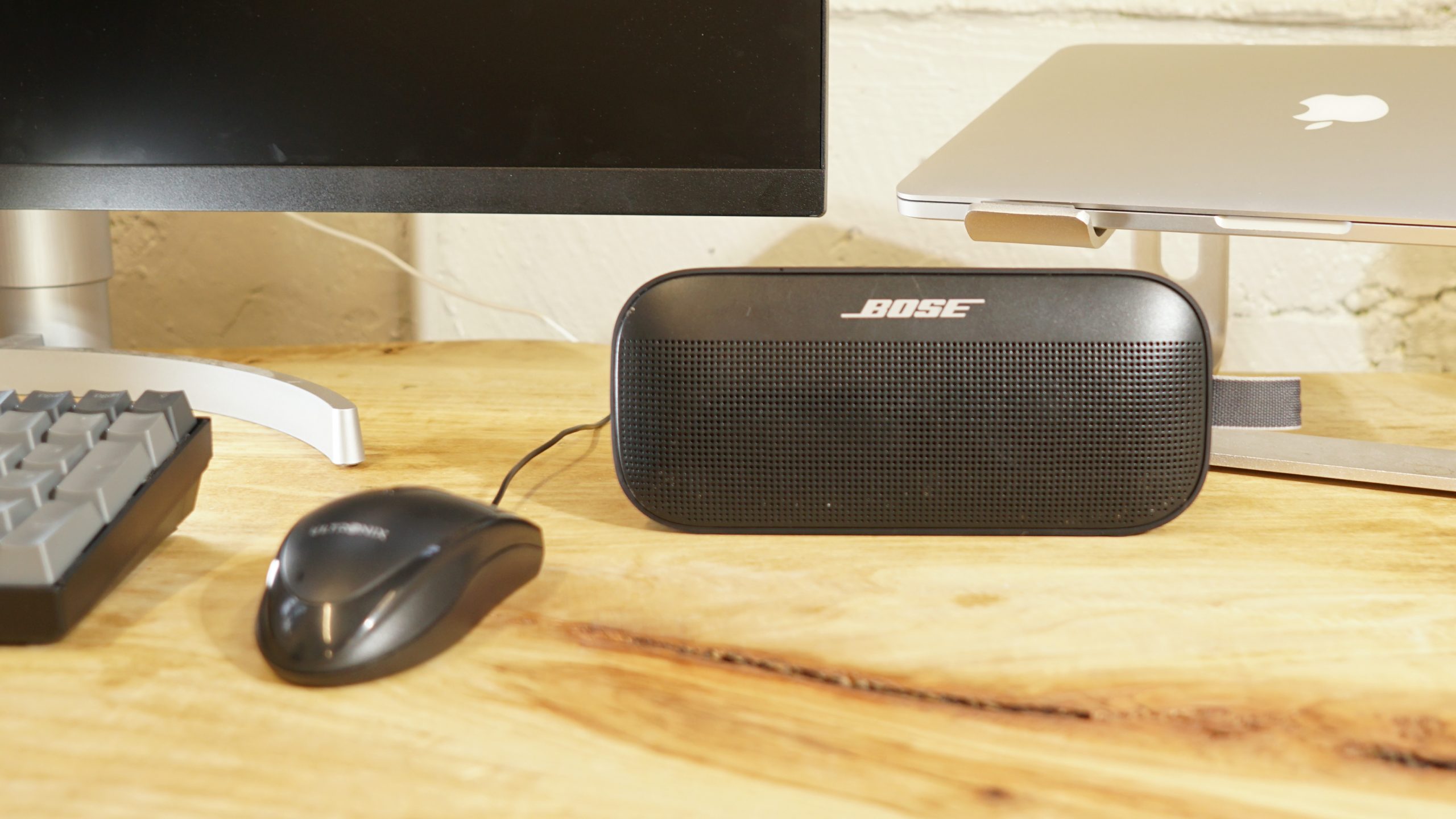 A photo of the Bose SoundLink Flex Bluetooth speaker sitting on a desk next to a monitor and laptop visible in the background and a mouse and keyboard to the left.