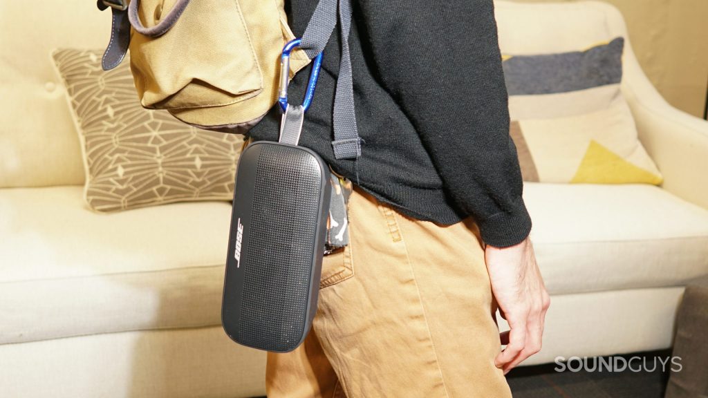 A black Bose SoundLink Flex bluetooth speaker hanging from a clip attached to a person's backpack near their right hip.