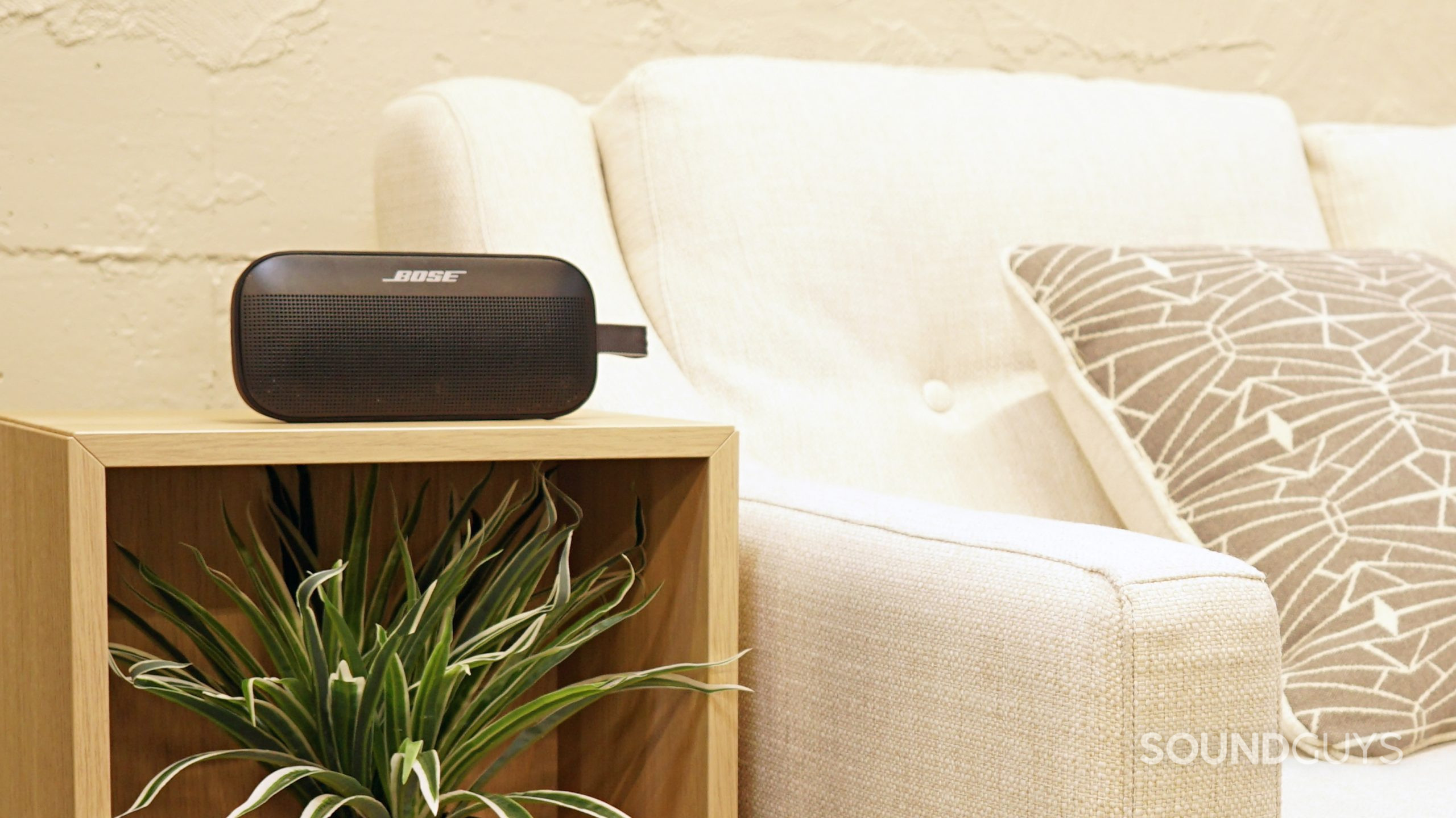 A photo of the Bose SoundLink Flex Bluetooth speaker sitting on a wooden box above a plant next to a white sofa.