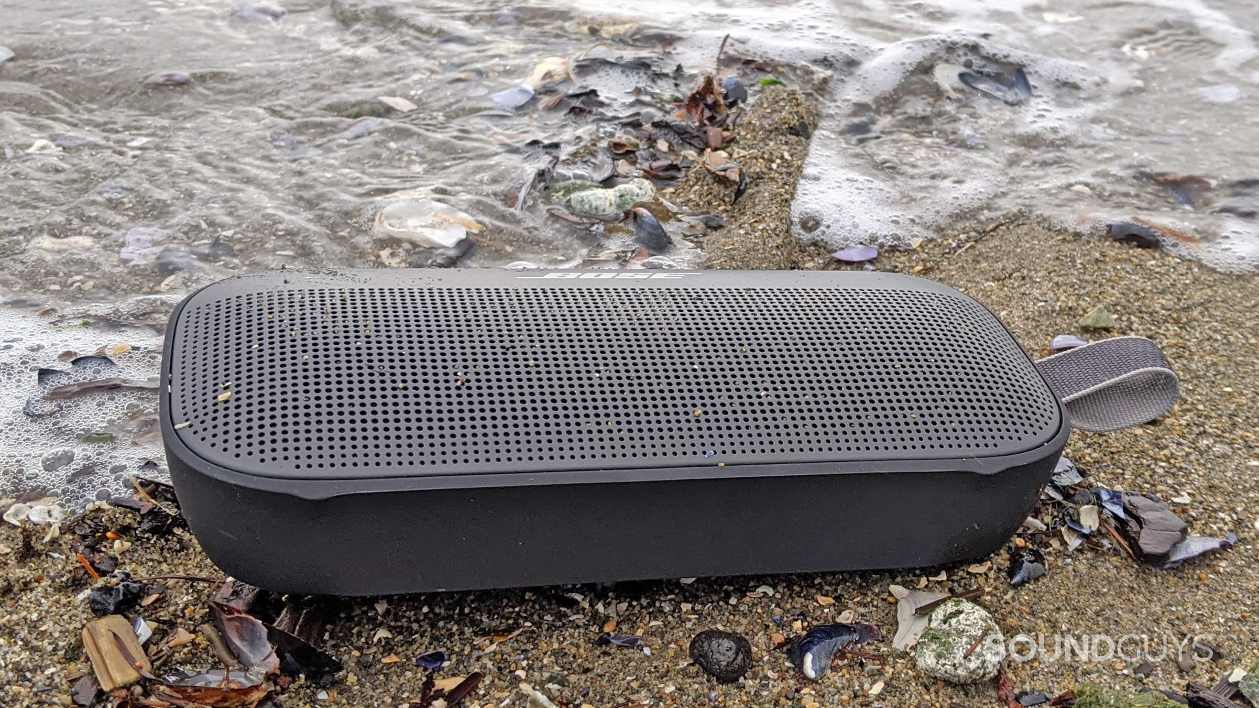 A photo of the Bose SoundLink Flex Bluetooth speaker lying face-up on the beach next to the ocean surrounded by shells and debris.