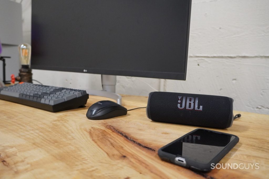 A JBL Flip 6 Bluetooth speaker sitting on a wooden desk next to a smartphone at its right and a monitor, keyboard, and mouse to its left.