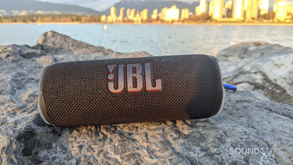 A JBL Flip 6 Bluetooth speaker sitting on a rocky surface with a sunny city skyline viisble in the distance.