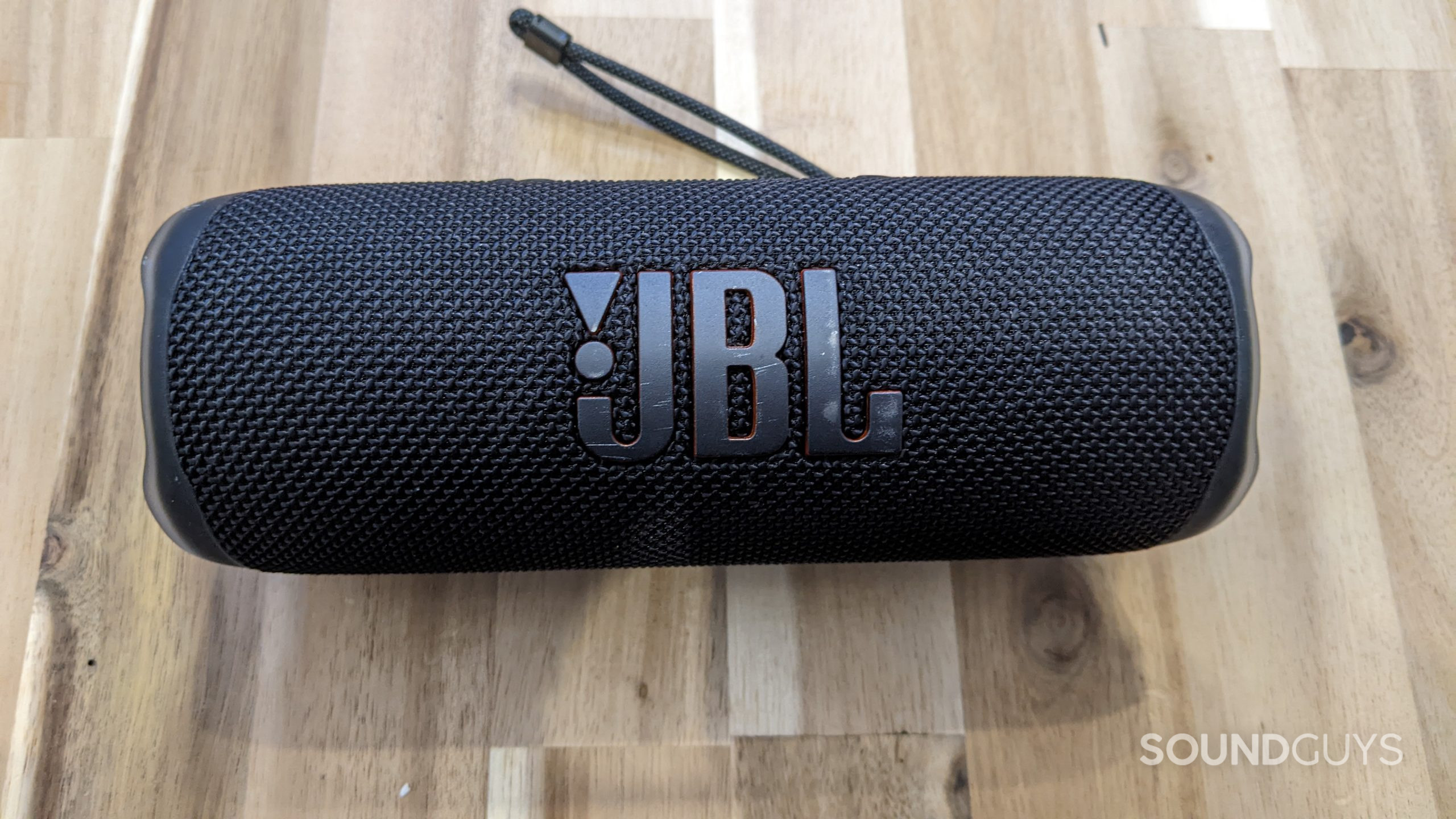 A JBL Flip 6 sittin on a table with the large &quot;JBL&quot; logo visible and the carrying strap seen in the background.
