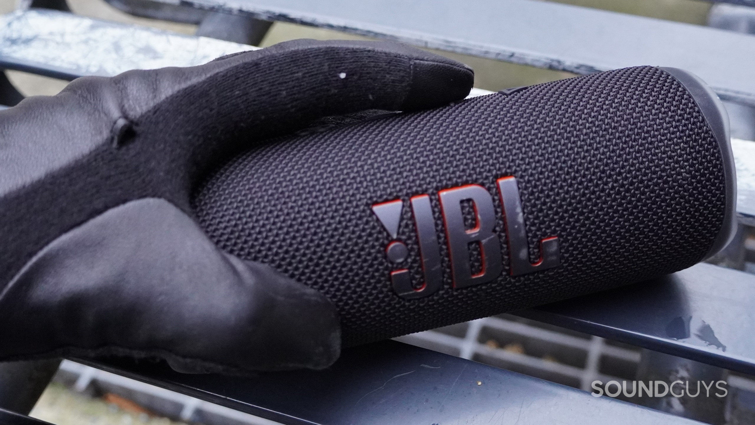 A hand wearing a black leather glove presses a button on the JBL Flip 6 Bluetooth speaker sitting on a metal bench.