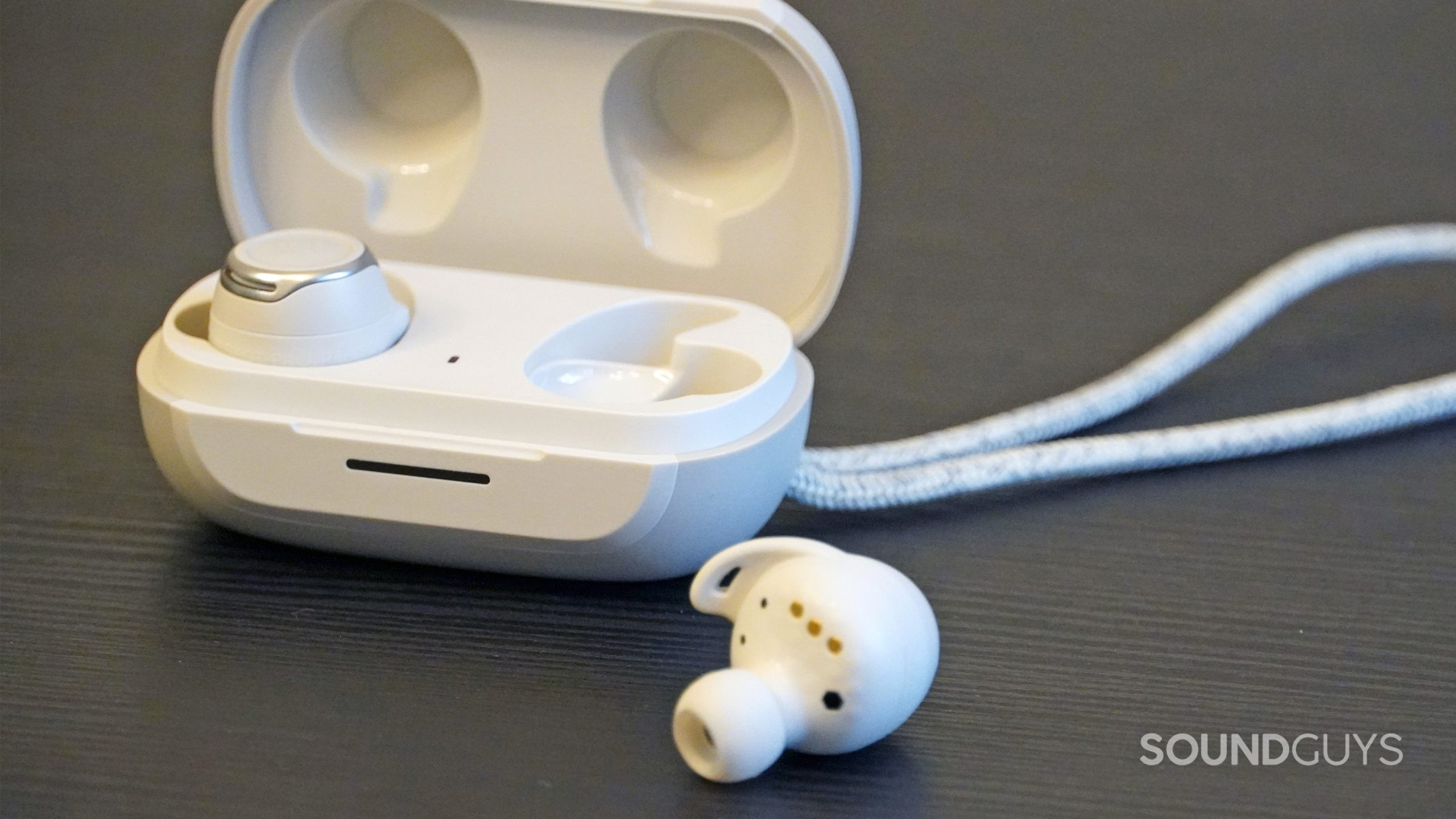 One JBL Reflect Flow Pro earbud outside of the case while the other is inside of the case behind it.