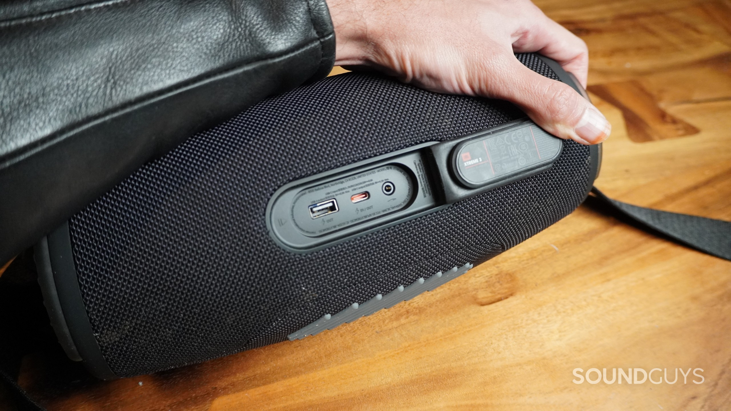 The JBL Xtreme 3 Bluetooth speaker showing its back flap being held open by a hand exposing the ports.