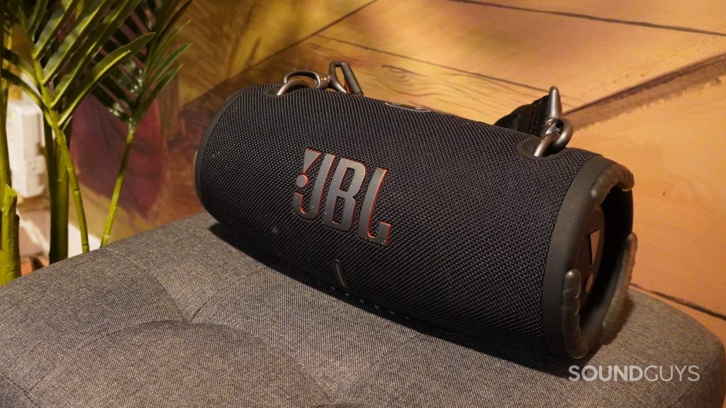 The JBL Xtreme 3 Bluetooth speaker sitting on a cushion by a fake potted plant and wall.