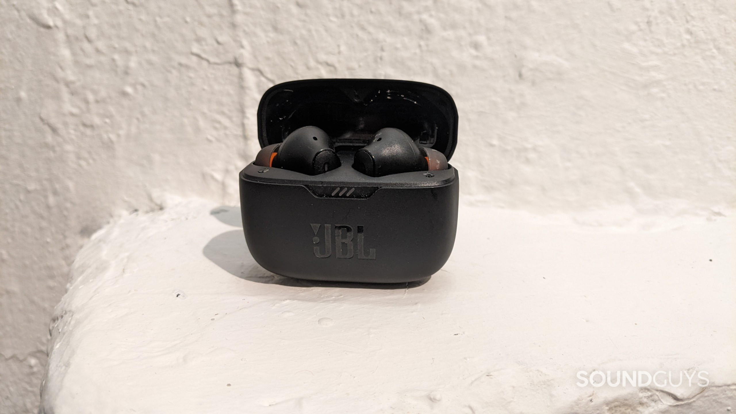 The JBL Tunes 230NC TWS earbuds in the charging case which is upright because it is carefully resting on a curved white surface.