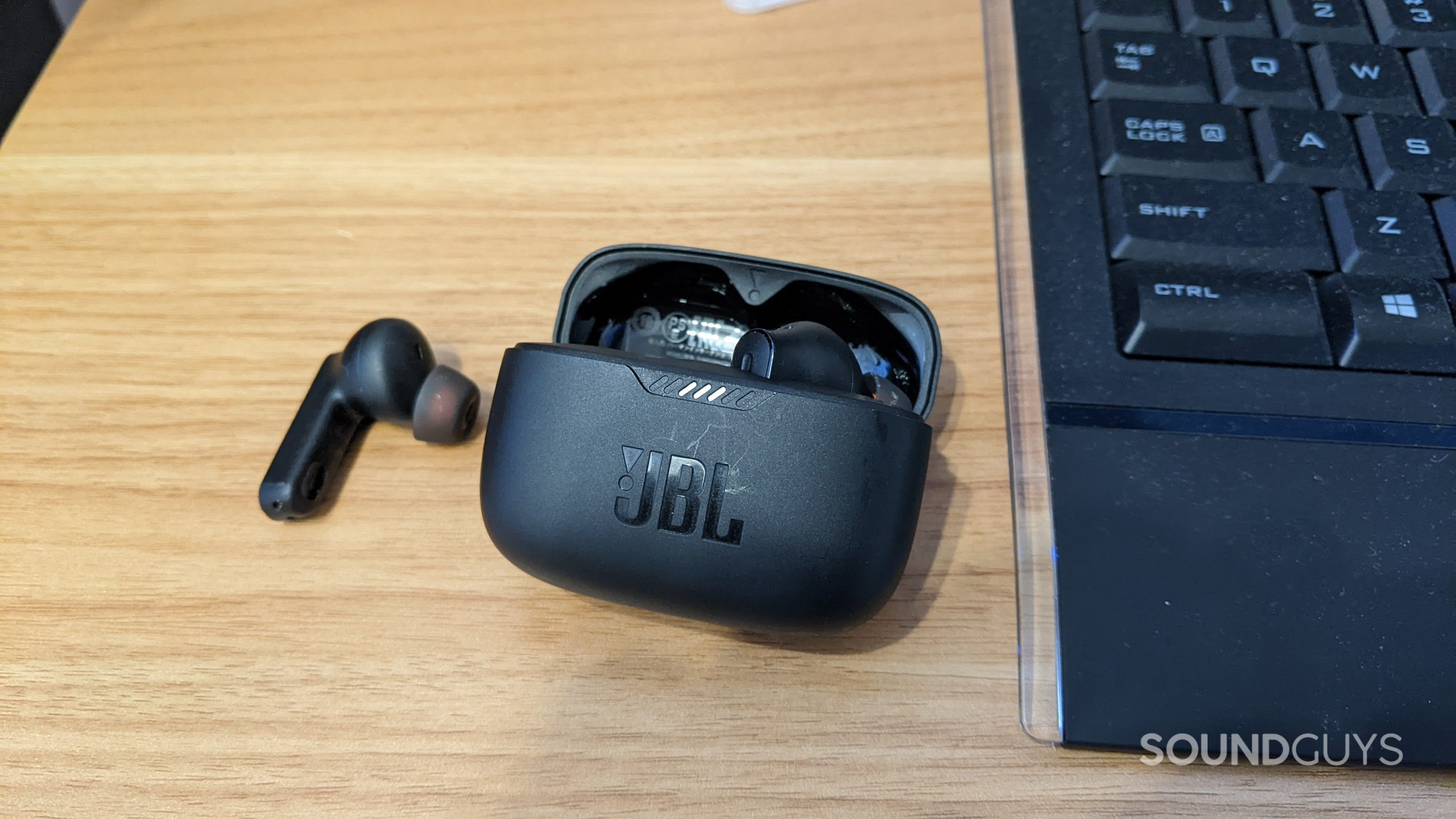 The JBL Tune 230NC TWS charging case lying next to a keyboard on a desk with the right earbud inside of it and the left earbud lying next to it.