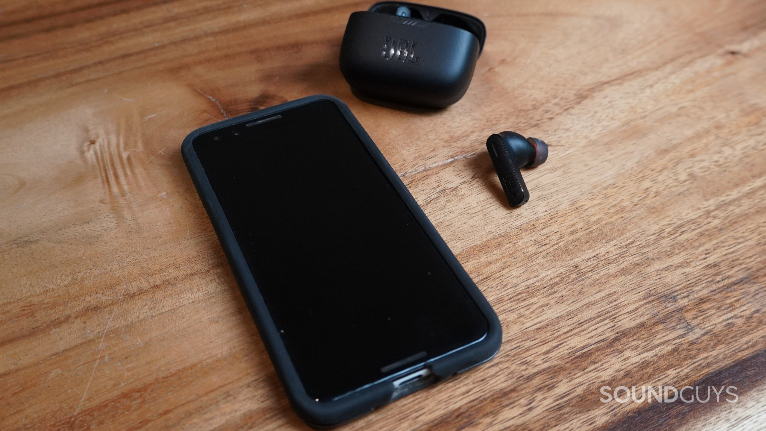 The JBL Tunes 230NC TWS earbuds shown next to a smartphone and the charging case on a wooden table.